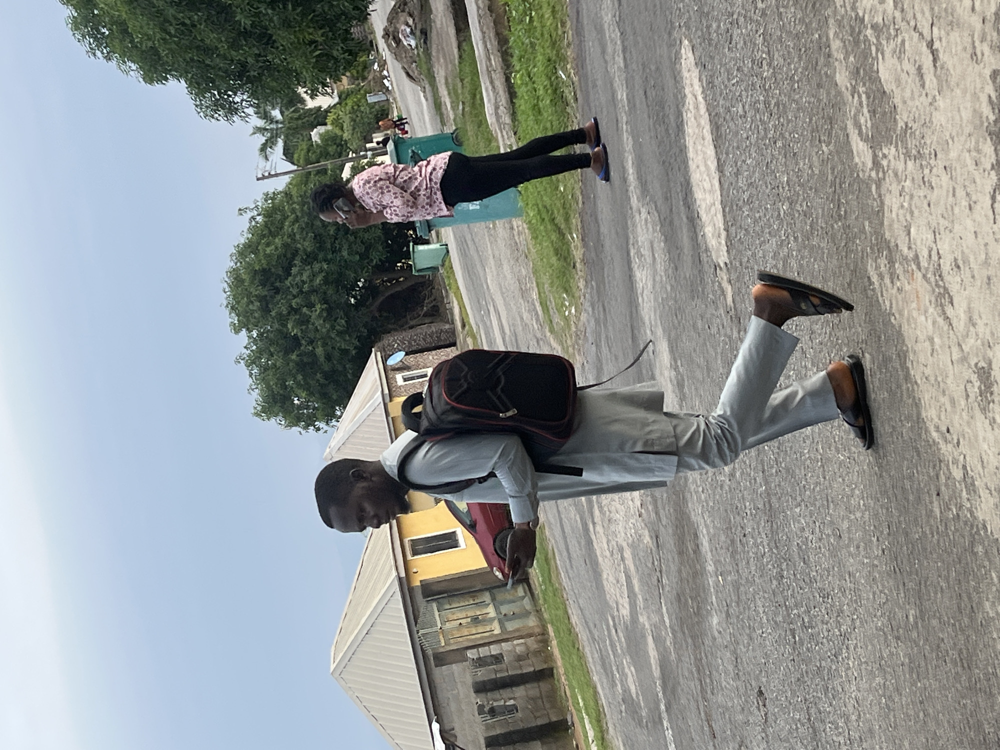 In an outdoor scene in Abuja, Nigeria, a Black man walks on an asphalt road, facing left, wearing a light blue long-sleeved tunic and matching pants, with a black and red backpack slung over his right shoulder. He holds a smartphone in his left hand. His feet are bare in dark sandals. In the background, a Black woman stands on the right side of the road, facing left and talking on her phone. She wears a pink floral long-sleeved shirt and black leggings, with blue and brown sandals. She is positioned in front of two large green trash bins and surrounded by green trees. Behind the man, a light yellow building with a silver corrugated roof is visible, adjacent to a grey brick structure with a gate and a red car parked in front. The sky is overcast.