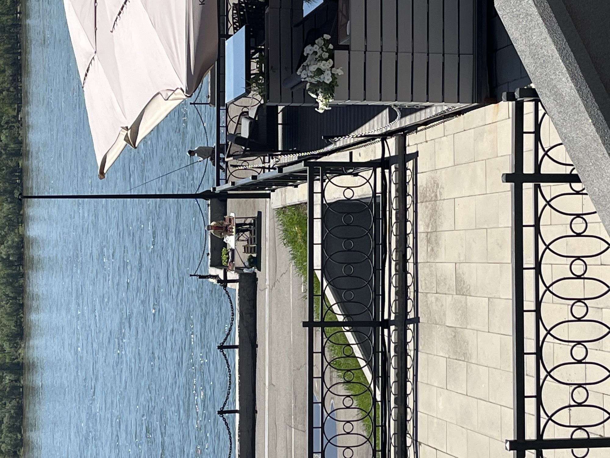 The image captures an outdoor cafe or restaurant terrace overlooking a wide body of water, likely a river or lake, on a bright, sunny day in Krasnoyarsk, Russia. The scene is illuminated by strong sunlight, creating clear reflections on the water and distinct shadows, suggesting it is midday or early afternoon.
In the foreground, a black metal railing with an ornate design of interconnected ovals and circles runs parallel to the viewer. This railing separates a paved walkway or path from the cafe area. To the right of this railing, a building facade constructed from light-colored bricks is visible, transitioning to a modern section with dark grey paneling further back.
To the left of the foreground railing, a paved asphalt path with white markings is present, alongside a narrow strip of grass. Beyond this, a lower black railing with draped chains borders the vast expanse of shimmering blue water. The distant bank across the water is lined with green trees.
On the cafe terrace, a large, light-colored umbrella or parasol provides shade in the upper left portion of the image. The partial text "KO" is visible on the umbrella. Beneath it, a person wearing a light-colored top is seated at a small table, facing the water. Another person can be faintly seen further back on the terrace. The terrace itself is furnished with additional outdoor seating. A decorative cluster of white flowers in a planter adds to the ambiance.
The overall atmosphere is one of peaceful relaxation, offering a scenic spot to enjoy the outdoors.