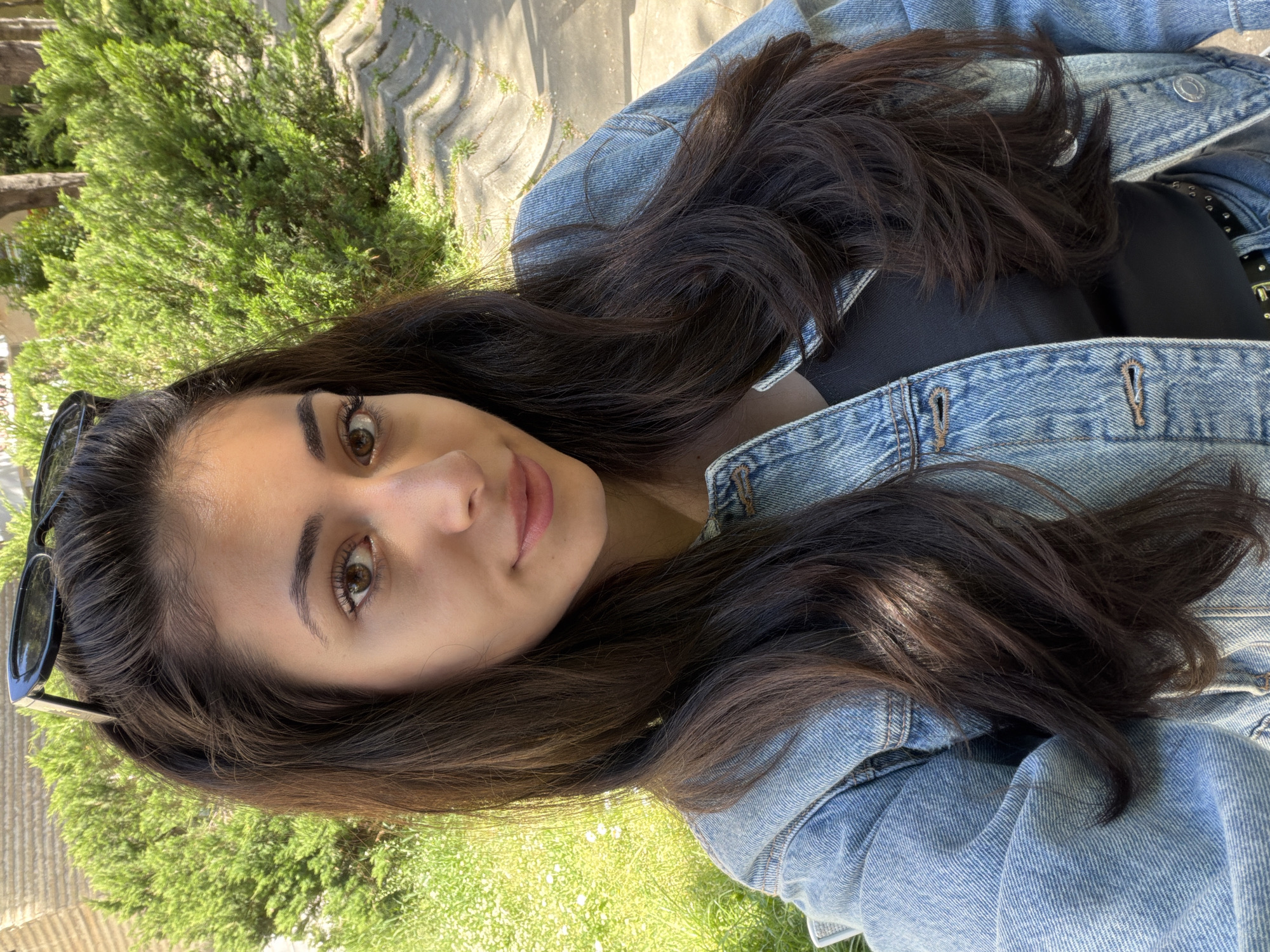 A young woman with long dark hair and brown eyes is taking a selfie outdoors. She is wearing a denim jacket and a black top. Sunglasses are perched on her head. The background consists of greenery, including bushes and grass. There is also a paved area with a pattern of interlocking shapes. The location may be a park or garden in the city of Blagoevgrad, Bulgaria, given the presence of vegetation and paving typical of urban green spaces.
