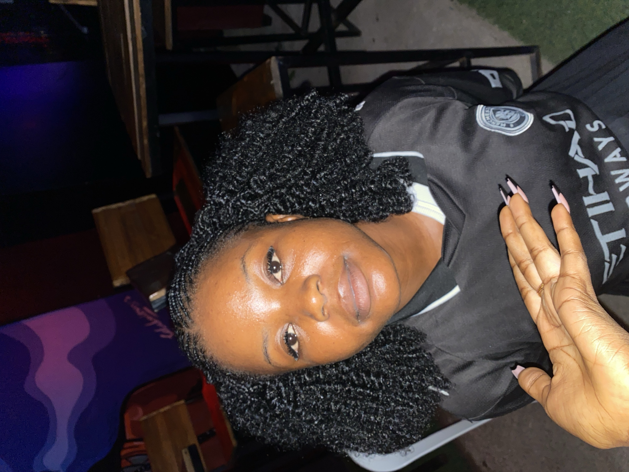 A medium shot captures a young Black woman from the chest up, posed indoors in a dimly lit setting that appears to be a bar or restaurant. She is seated and facing the camera, her skin appearing dewy. Her dark, braided hair is styled in two pigtails that frame her face. She is wearing a black jersey with a white collar and a circular crest on the left side of her chest. A hand, presumably belonging to another person, is placed on her chest, with manicured fingernails visible. The background includes wooden tables and chairs, and a portion of a wall decorated with wavy, colorful abstract art in shades of purple and pink.