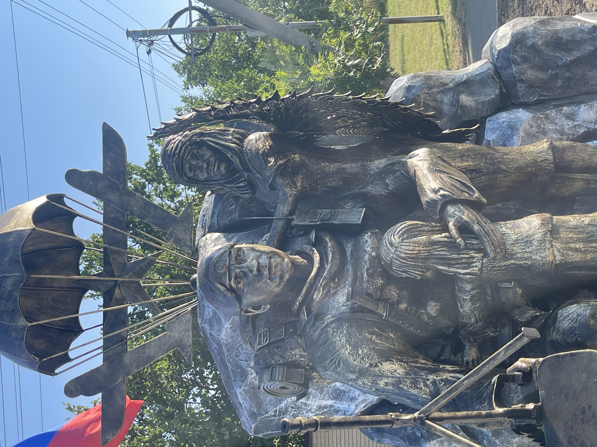 This is a frontal, medium-shot photograph of a bronze monument located in Budyonnovsk, Russia, depicting a paratrooper holding a child. The monument is situated outdoors with a clear blue sky and green trees in the background, along with utility poles and wires.
The central figure is a male paratrooper, rendered in bronze. He is wearing a beret, a tactical vest, and a backpack with a rolled sleeping bag attached. A rifle is strapped to his side. He is looking forward with a stern expression. He is embracing a young child, also depicted in bronze, who is being held protectively. The child has long hair and appears to be looking down.
Behind the figures, the monument features a large, rough-textured bronze rock formation. Emerging from behind the paratrooper and child is a second figure, a woman with a scarf wrapped around her head, also in bronze. She is holding a laurel wreath.
Above the figures, a large parachute canopy is depicted, with rigging lines extending downwards. Underneath the parachute, there is a stylized bronze airplane with a star emblem on its wing. To the left of the monument, a Russian flag (tricolor of white, blue, and red) is partially visible, attached to a flagpole.
The monument appears to be situated in a public space, possibly a park or a square, with a paved path and grass visible in the background. The overall scene conveys a sense of protection, heroism, and perhaps remembrance.