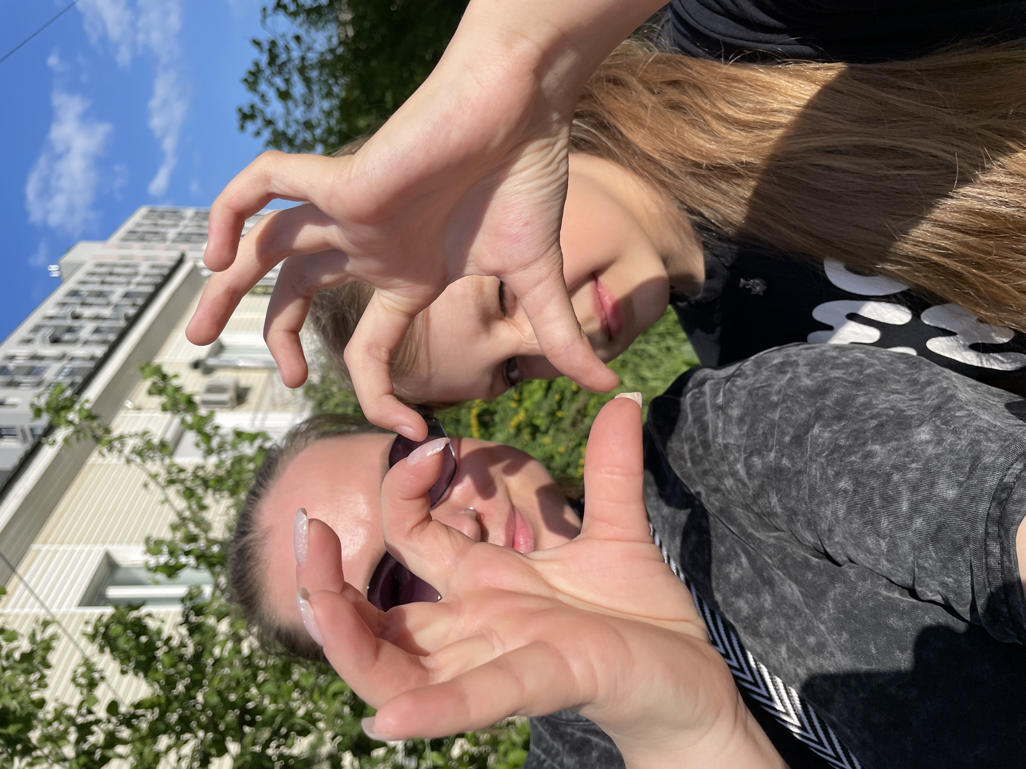 Two young women are posing for a selfie outdoors. The selfie is taken from a low angle, looking up at them. The faces of both women are partially obscured by their hands, which they are holding up in front of their faces. The woman in the foreground is wearing sunglasses and has her hair pulled back. The woman behind her has long, blonde hair. Behind the women, there is a building, some trees, and a blue sky with clouds. The location appears to be in the city of Izhevsk, Russia.
