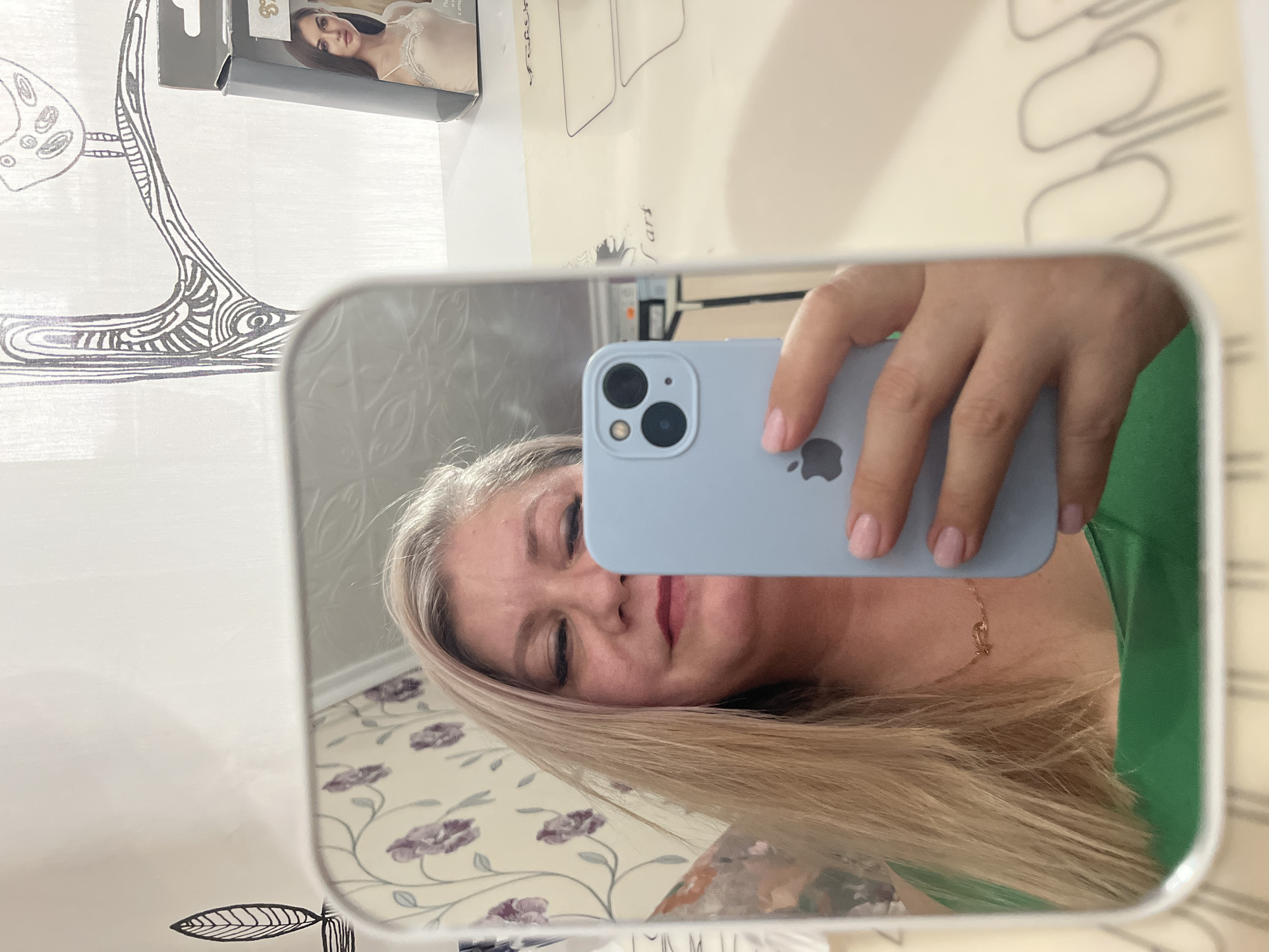 A woman is taking a selfie in a mirror. She is holding a light blue iPhone, presumably to take the picture. The reflection shows her face, light blonde hair, and a green shirt. A gold necklace is visible around her neck.
The background, also visible in the mirror's reflection, features a floral patterned wall and what appears to be a piece of furniture. Outside of the mirror, elements present include a box with a woman's image on it and a patterned curtain. The specific location within Izhevsk, Russia, cannot be determined from the given image.