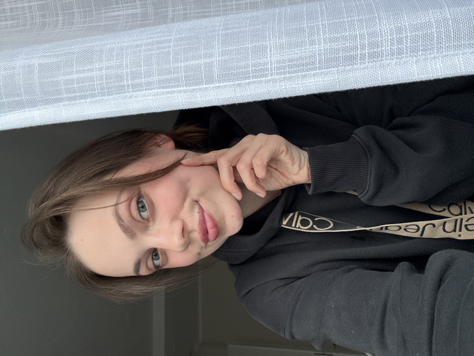 A young adult female is depicted in a close-up, chest-up indoor portrait, presumed to be taken within the city of Scafati, Italy. She has brown hair, blue eyes, and fair skin, with a slight smile and a visible dimple on her right cheek. Her right hand is positioned with fingers touching her right jawline. She is wearing a black hooded sweatshirt with a beige drawstring displaying the repeated text "Calvin Klein Jeans". The background to her left is a light gray, blurred surface. To her right, a white, semi-transparent curtain with a discernible grid pattern partially frames the scene.