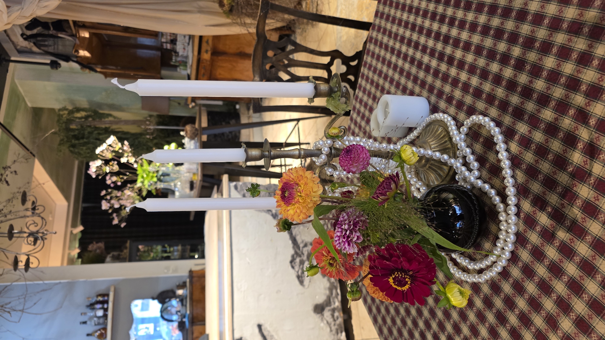 The image depicts a table setting indoors, likely in a restaurant or home, with a checkered tablecloth. On the table, a brass candelabra holds three white taper candles. A bouquet of vibrant flowers, including dahlias in shades of orange, yellow, pink, and deep red, is arranged in a dark, glossy vase. A strand of faux pearls is draped around the candelabra and vase. To the right of the candelabra, two white pillar candles are present. In the background, a large window reveals a blurred outdoor scene, and further back, shelves with bottles and decorative items are visible. The overall ambiance suggests a decorated interior space. There are no people present in the image. The image does not contain any specific details that would allow for identification of its location in Riga, Latvia.