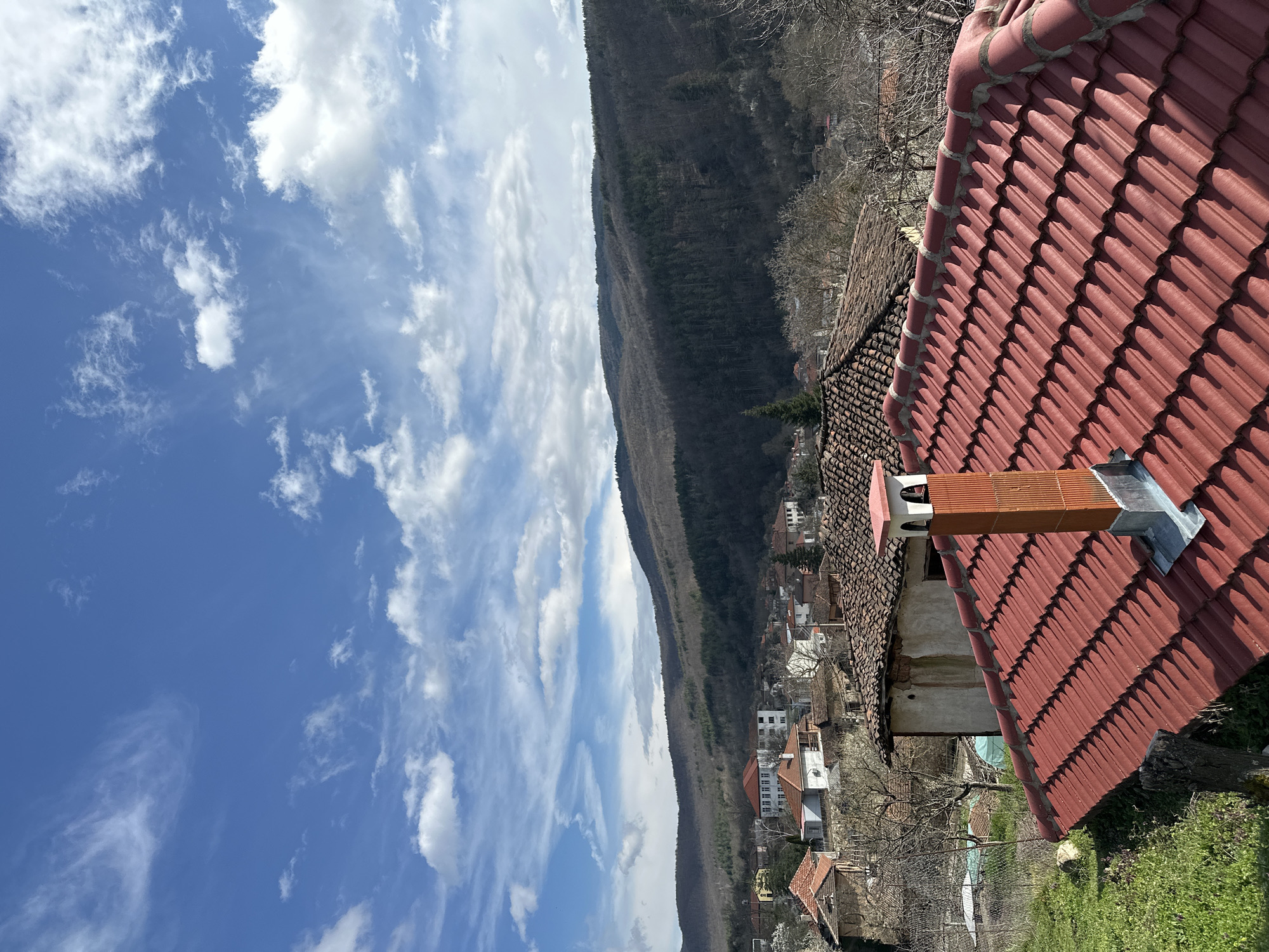 The scene, set in Mrachenik, Bulgaria, presents a high-angle view, potentially from a rooftop, capturing the landscape and parts of the village. A predominantly red tiled roof occupies the foreground, with a chimney extending upwards. Beyond the rooftops, a row of buildings with red-tiled roofs lines a valley, giving a sense of the settlement. A densely wooded mountain forms a prominent backdrop, its slopes ascending steeply. The sky is mostly blue, scattered with wispy and puffy clouds. The image is vertically oriented. There are no people visible in the frame.