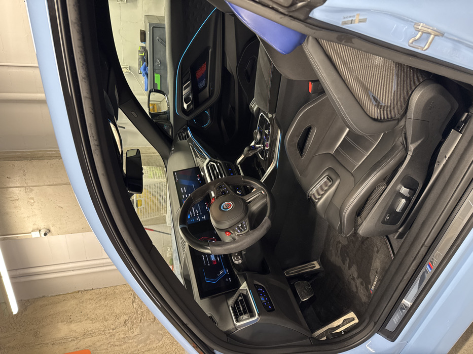 The image presents the interior of a light blue two-door coupe, viewed from an open driver's side door, looking towards the passenger side. The interior features black leather and Alcantara sports seats with visible carbon fiber accents on the seat sides and bolsters. The driver's seat includes electric adjustment controls. The steering wheel is flat-bottomed, wrapped in Alcantara, adorned with an "ALPINA" logo badge, and features red "M" buttons and carbon fiber trim.
The dashboard incorporates two illuminated digital screens: an instrument cluster displaying vehicle data and a central infotainment display. Blue ambient lighting runs along the dashboard, air vents, and door panels. The passenger door panel also displays a section with red and blue striped accents. The center console includes an automatic gear selector, an iDrive controller, and carbon fiber trim. Black floor mats are present on the driver's side, and an "M" logo is visible on the illuminated door sill. No individuals are present in the vehicle.
The car is located indoors, within an apparent garage or workshop environment. The background shows an unfinished or concrete wall with horizontal indentations. Fluorescent strip lighting is mounted on the ceiling, and a white security camera is affixed to the wall above the windshield. Through the car's windows, a white plastic basket, a dark green metal toolbox topped with blue and yellow items, and a red and white striped object are partially visible against the background wall. No specific visual information within the image identifies a particular location in Warsaw, Poland.