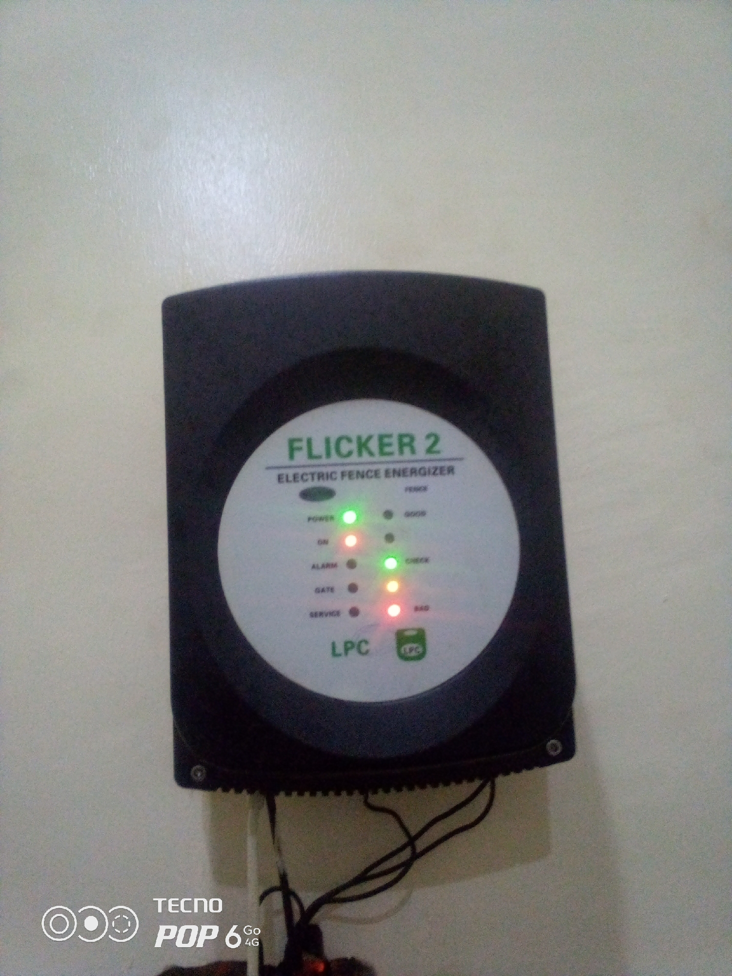 The image shows an electric fence energizer, specifically a "FLICKER 2 ELECTRIC FENCE ENERGIZER", mounted on a lightly textured, off-white wall. The device is black and circular in the center, with various indicator lights. A green light is illuminated next to "POWER" and "CHECK". A red light is illuminated next to "ON" and "BAD". The device also has labels for "ALARM", "GATE", "SERVICE", and "FENCE". Below the central display, the letters "LPC" are visible, along with a small green circular logo that also reads "LPC". The bottom of the image shows some black electrical wires connected to the device and a watermark that says "TECNO POP 6 Go 4G". The lighting in the image is dim, suggesting it was taken indoors or at night. There are no people visible in the image. The device appears to be in working condition, with multiple lights illuminated. The background is a plain wall, with no visible windows or other furnishings. The scene does not provide any specific geographical clues that would indicate its location within Howayi, Nigeria, beyond what might be inferred from the brand watermark.