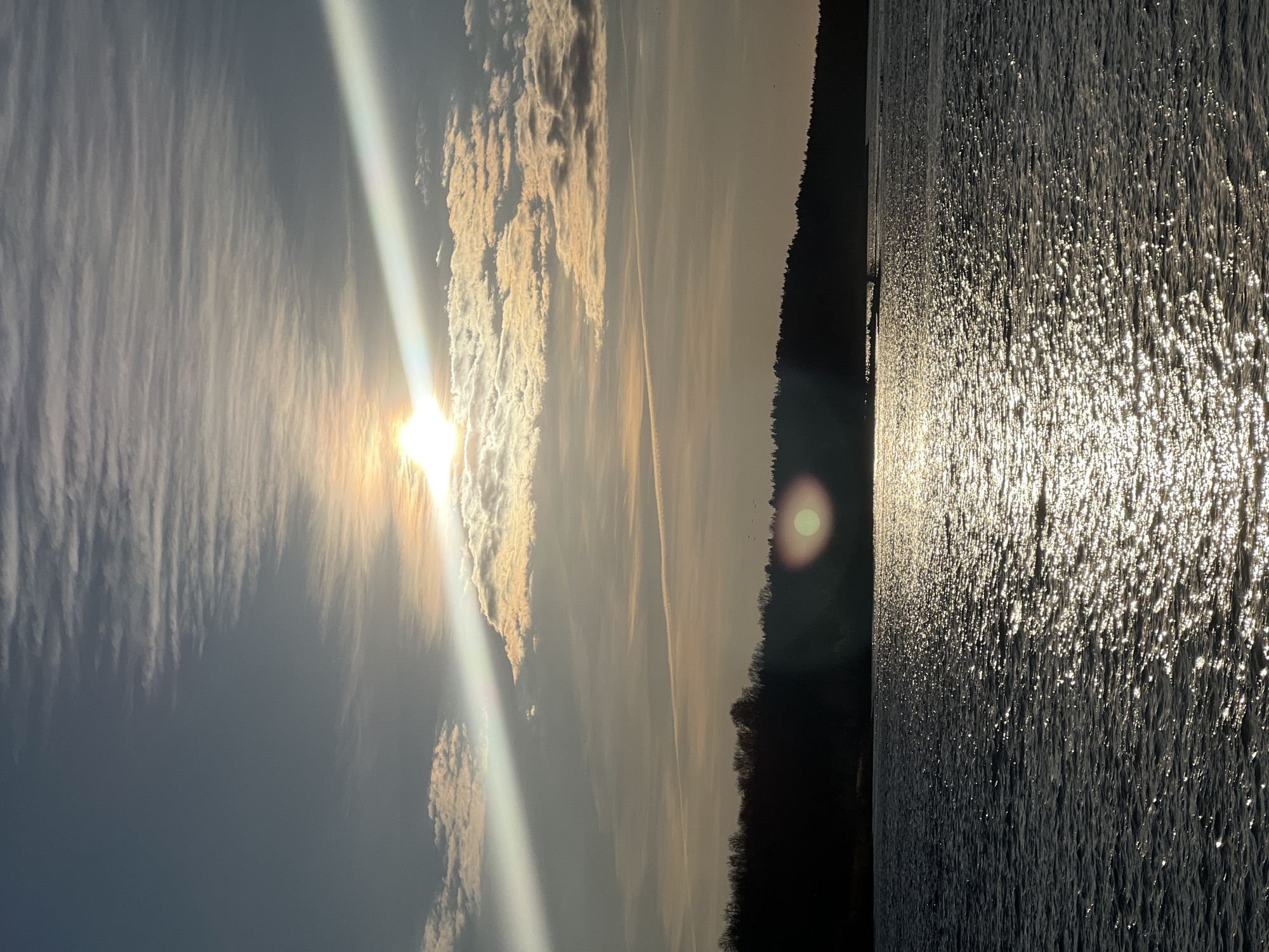 The image depicts a waterfront scene. The sun is bright and appears to be low in the sky, casting a strong reflection on the rippled water's surface. The sky features varied cloud formations, some dense and others wispy, with the sun's rays shining through. A dark shoreline or treeline is visible in the distance. No people are discernible in the image. The location in the city of Riga, Latvia, is not determinable from the visual information provided.
