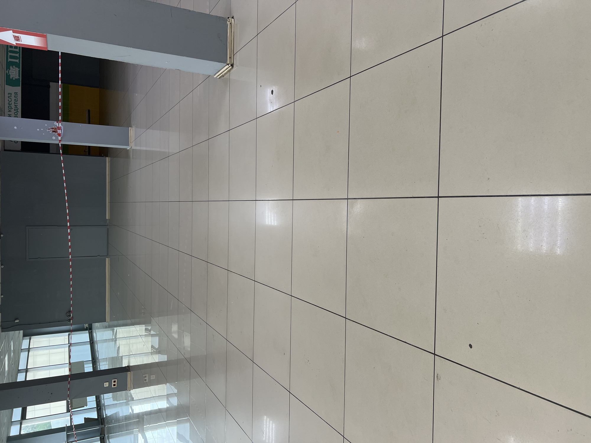An indoor scene depicts a wide, brightly lit, polished tile floor with a grid pattern. The floor is clean, with some minor scuff marks and a few dark spots. In the background, a gray wall with a red and white striped caution tape is visible. Several large, gray pillars stand on the floor, two of which are prominent in the foreground and middle ground. The pillars have reflective golden bases. To the left, large windows with a criss-cross design show a bright, reflective interior space. A radiator is visible behind the windows. To the right, a portion of a sign with green lettering and an arrow is visible. The overall environment appears to be a large, empty commercial or public space.