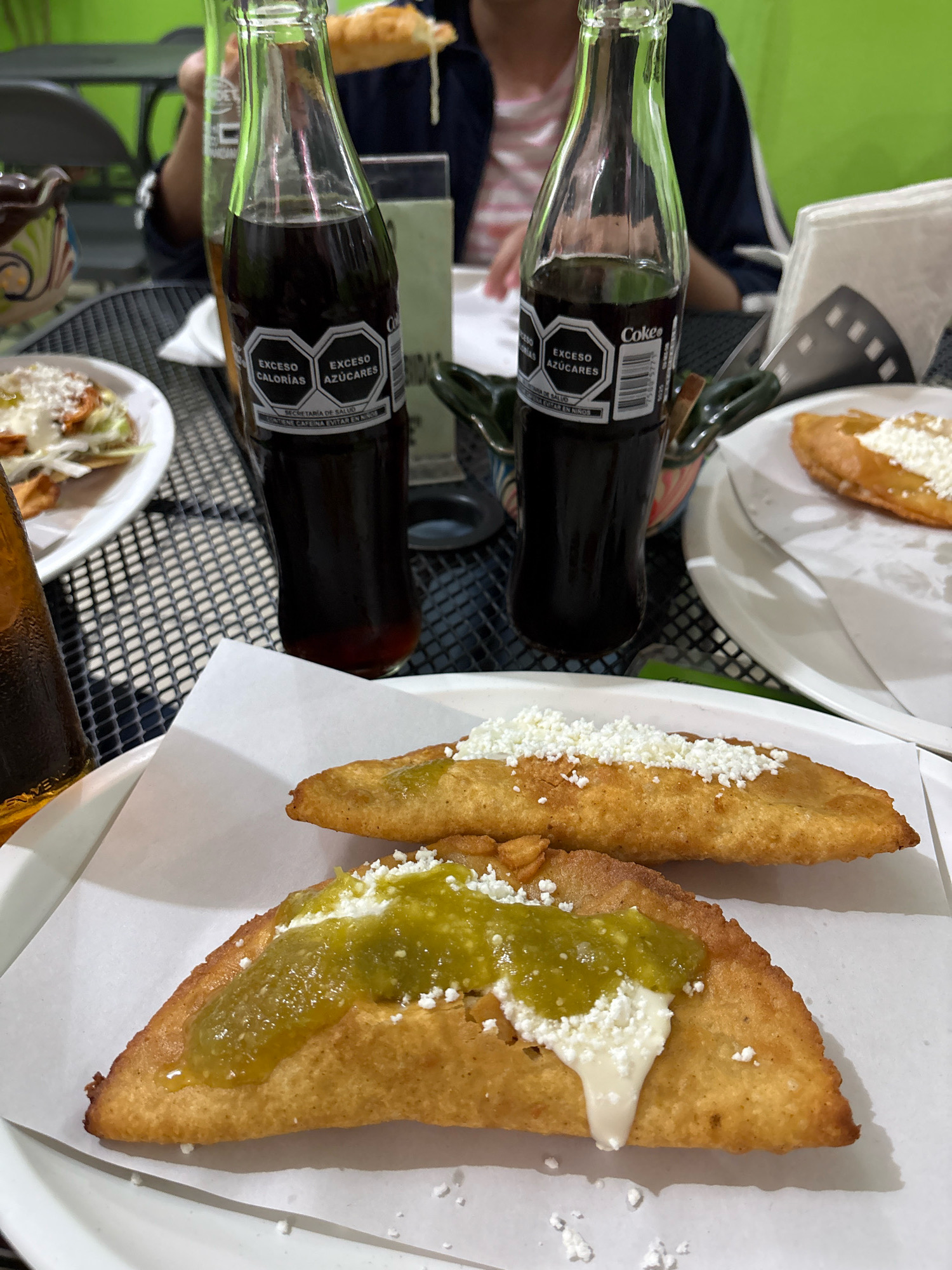 The image shows two fried pastries, possibly empanadas, topped with crumbled white cheese and a green sauce. They are served on white paper on a white plate. In the background, there are two glass bottles of Coca-Cola with warning labels about excess calories and sugar, typical of those sold in Mexico. A person's hand is visible in the upper left, holding a pastry. The scene appears to be set at an outdoor table with a dark, patterned surface. The overall setting suggests a casual dining establishment, likely a street food vendor or a small restaurant. The presence of Mexican-labeled Coca-Cola bottles and the style of food strongly suggest the location is Mexico. While not explicitly stated, the presence of palm trees and the characteristic bright green paint on the wall in the background are common elements found in the city of Cuernavaca, known as the "City of Eternal Spring."