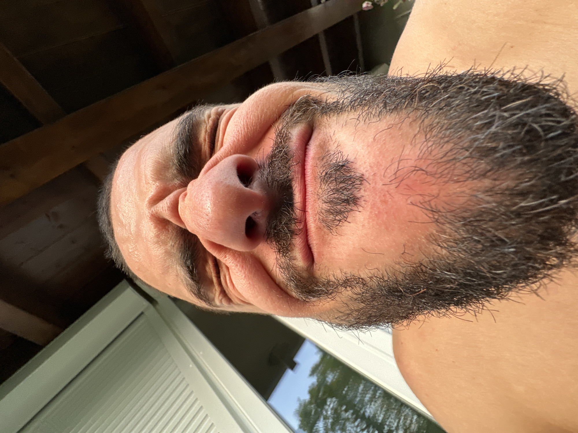A man with a full beard and dark hair is taking a close-up, low-angle selfie outdoors. He is squinting his eyes and making a funny face, with his nose wrinkled and his lips pursed. The background shows wooden beams, a window with light-colored blinds, and some greenery. The image appears to be taken in a backyard or patio area. There is no indication of the city of Garmisch-Partenkirchen, Germany in this image.