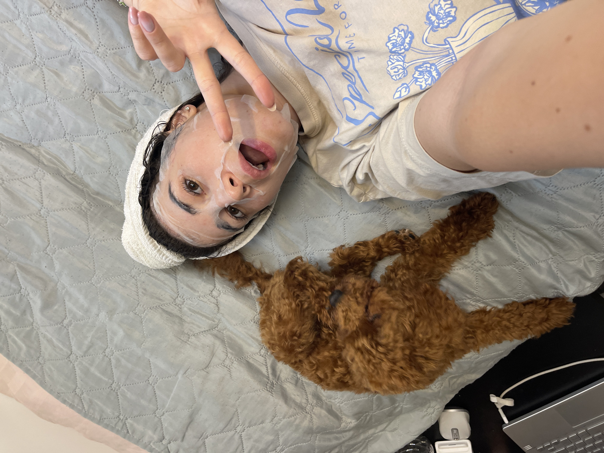 The image shows a young woman lying on a bed with a sheet mask on her face and a white towel wrapped around her head. She is looking at the camera and holding up two fingers in a peace sign. Beside her lies a small, brown poodle. A laptop and a small white electronic device are visible on the left side of the image. The room appears to be a bedroom. There is no information in the image to determine the location in the city of Sofia, Bulgaria.