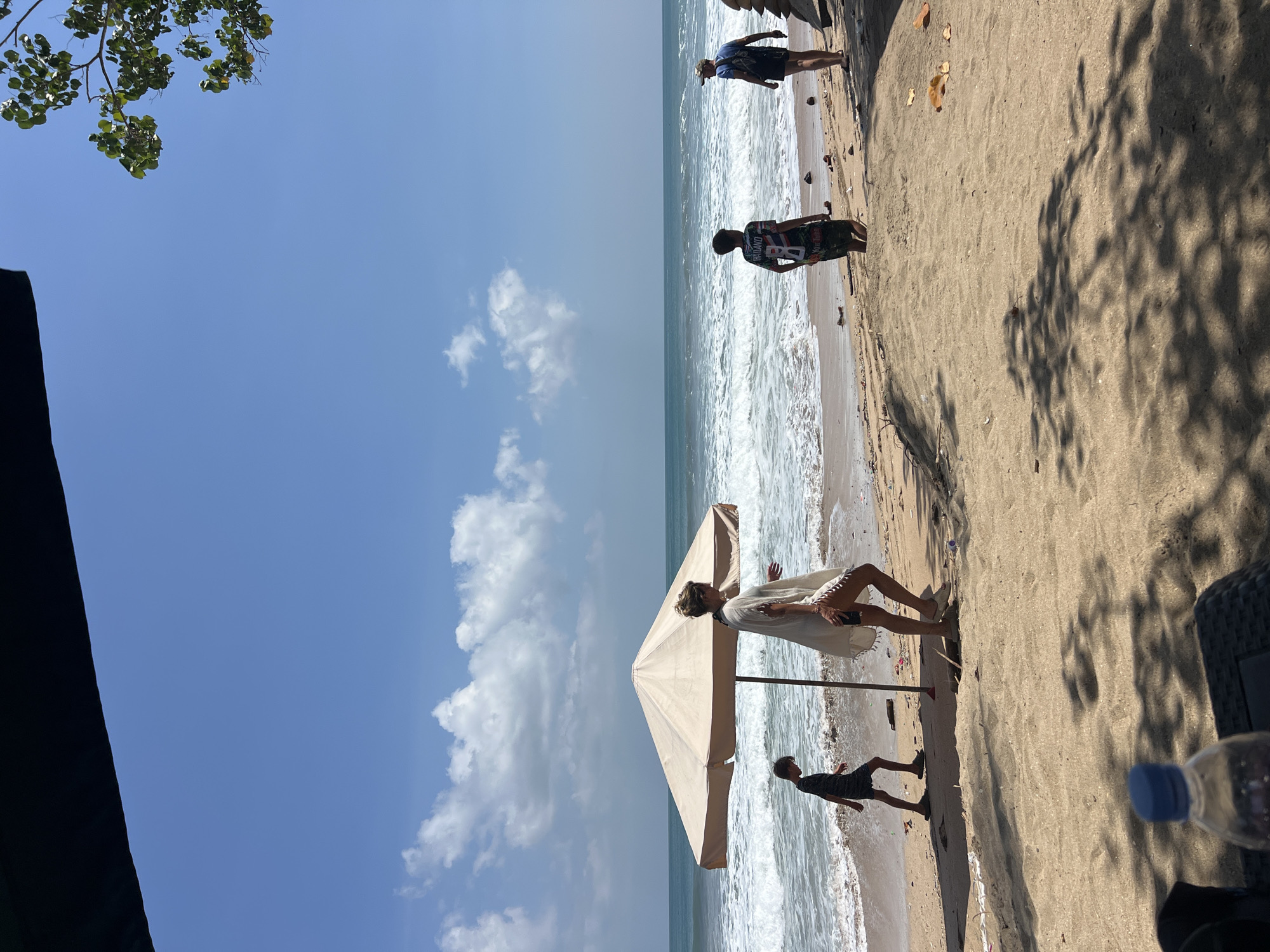 In Kuta, Indonesia, a beach scene unfolds under a clear blue sky with scattered white clouds. The ocean exhibits moderate waves, crashing onto a sandy shore. In the foreground, a large, tan umbrella provides shade, and a woman in a white cover-up walks away from it, her legs and feet visible. To her left, a young boy in a dark striped shirt and shorts walks along the water's edge. Further to the right, two other individuals, a boy wearing a colorful t-shirt with "THAILAND" and "You Tube" printed on it, and a man in a blue t-shirt and shorts with a backpack, stand near the water. Debris, including plastic bottles and leaves, is scattered across the sand. The scene is framed by the dark edge of an awning at the top and shadows of foliage cast on the sand in the lower portion.