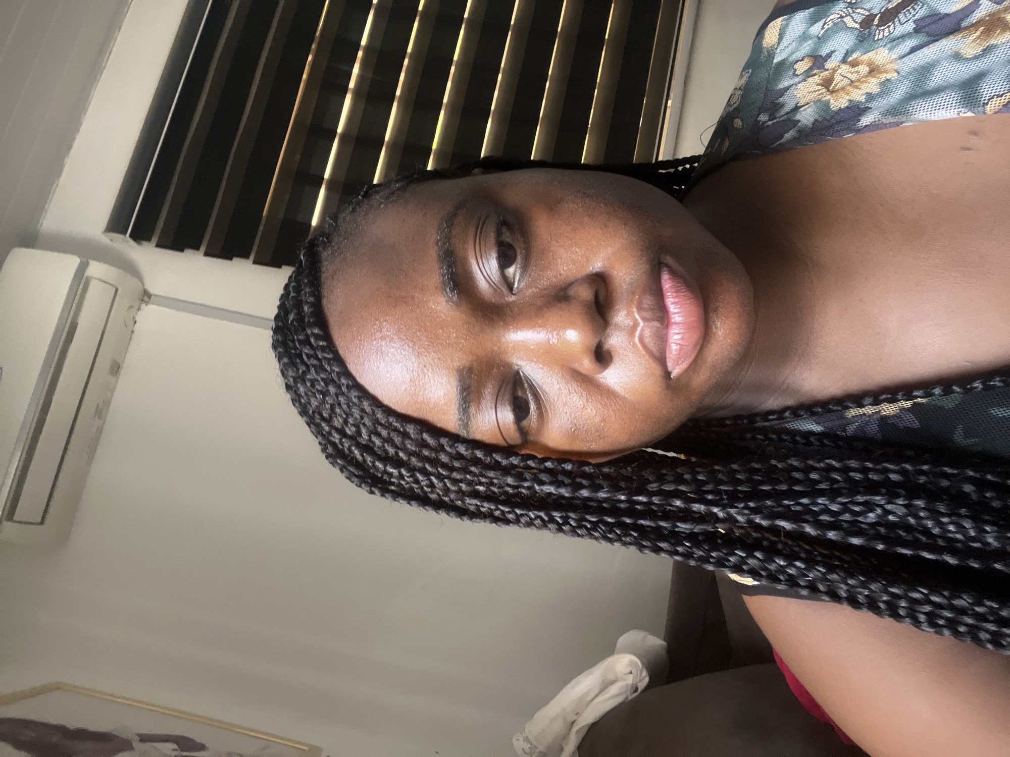 A close-up, slightly low-angle selfie features a young Black woman looking directly at the camera. Her face is well-lit, showcasing her smooth, dark complexion with a natural sheen, especially on her forehead, nose, and full, pinkish lips. Her dark eyes are open, and she has visible eyebrows. Her hair is styled in numerous neat, dark box braids that frame her face and extend over her shoulders.
She is wearing a top with a green, yellow, and black floral or patterned design, visible on her upper chest and shoulder. A glimpse of a red garment, possibly shorts or underwear, can be seen near her lower body.
The setting appears to be indoors during the daytime. Natural light filters through horizontal window blinds, casting alternating stripes of light and shadow on the wall in the background. To the left of the woman's head, a white wall-mounted air conditioning unit is visible. Below the blinds and to the right, a dark piece of furniture, possibly a couch, is present, with a crumpled white cloth or towel resting on it. A framed picture with a gold border is partially visible in the bottom left corner of the scene. The overall ambiance is bright and casual. There is no visible text in the image.