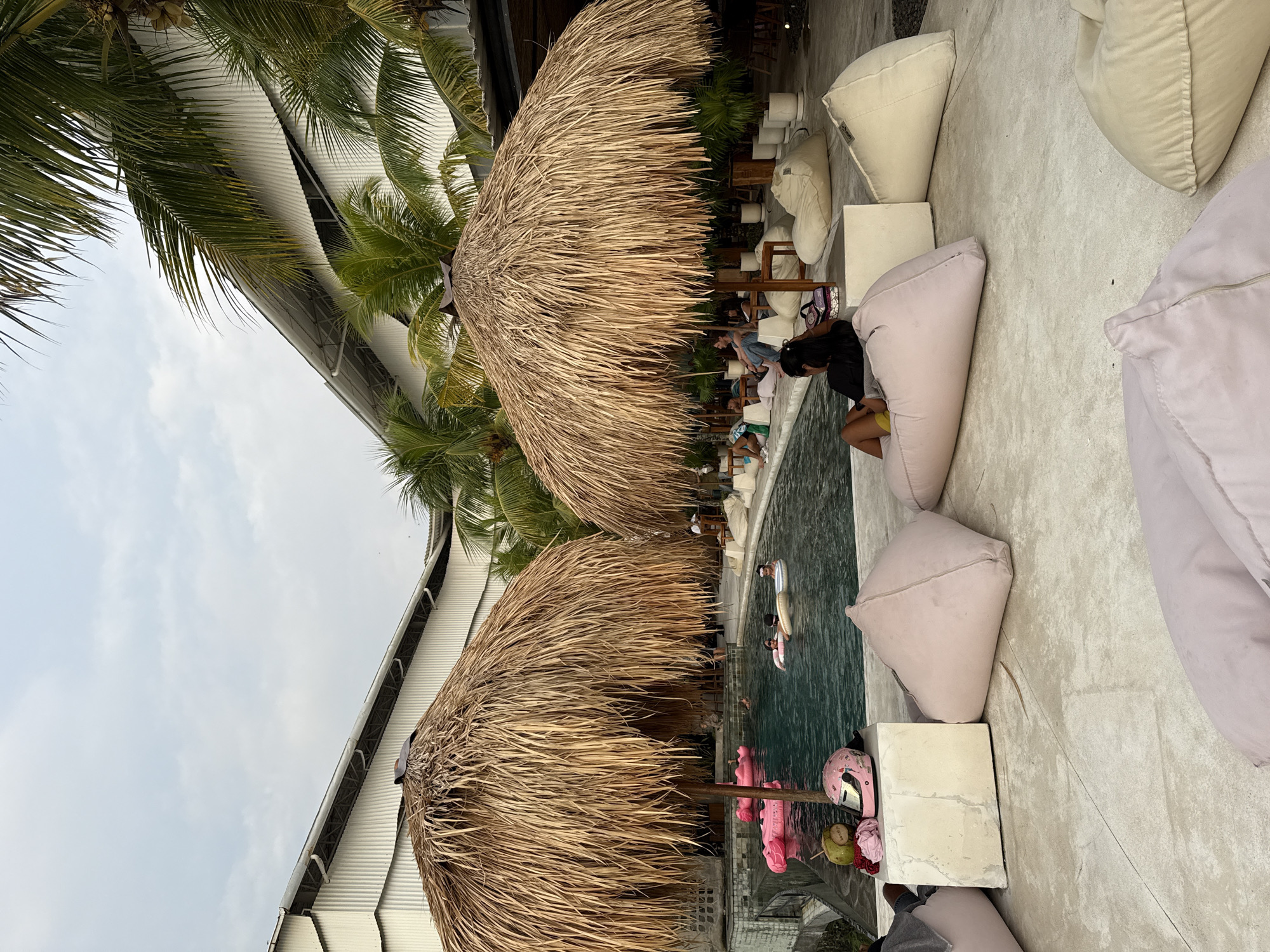 The image depicts a scene at a poolside bar/lounge in Canggu, Indonesia. Two thatched umbrellas dominate the foreground, providing shade over a partially visible pool area. Several people are swimming in the pool on floats, and others are seated at tables and chairs near the water's edge. Around the pool are beanbags and white cube seats. There is a corrugated metal roof to the left side of the scene, partially obscured by palm trees. The sky is overcast. A pink motorcycle helmet and a coconut drink are placed on top of one of the white cube seats.