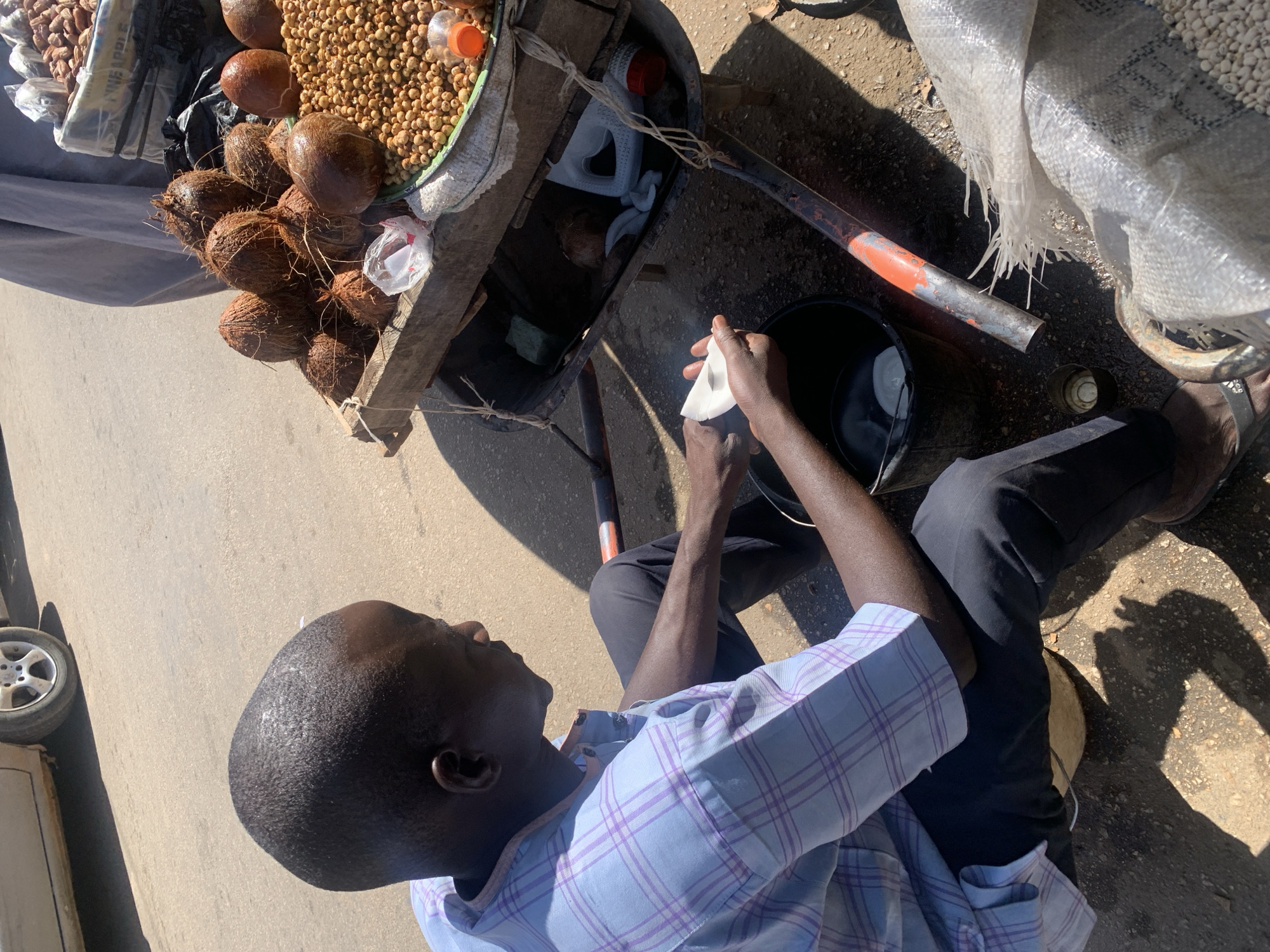 A man sits on the ground next to a makeshift cart. The cart contains coconuts, beans, and a bottle of juice. The man holds a coconut and uses a rag to wipe it. A black bucket sits next to the cart. The location appears to be an urban area, possibly Jos, Nigeria, based on the context. The ground is asphalt.