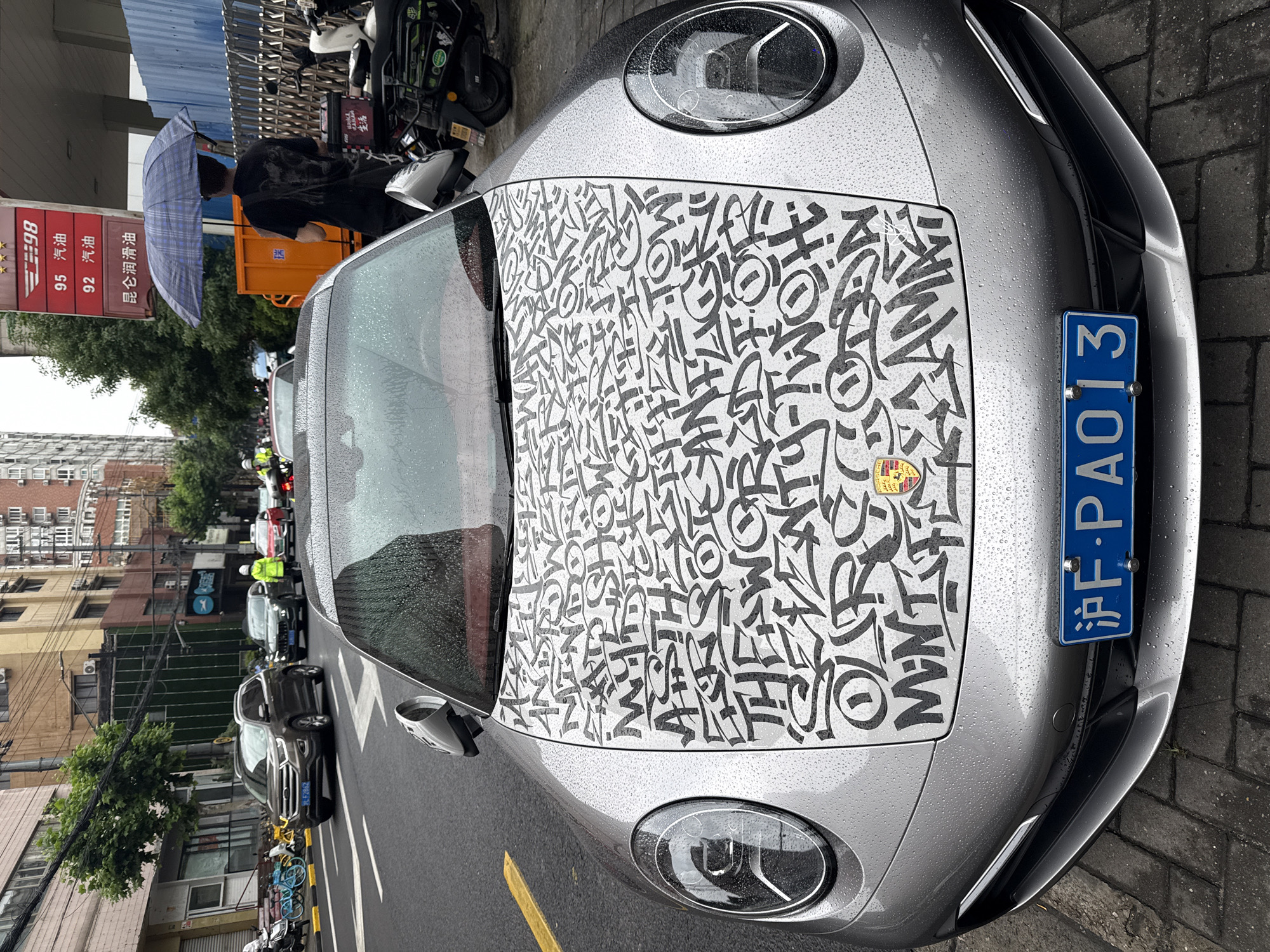 This urban street scene, likely in Jing'an District, China, is captured on an overcast day with recent rain, as indicated by visible water droplets on surfaces. The focal point is a silver Porsche sports car, possibly a 911, parked roadside. Its hood features a distinctive black-and-white graphic wrap, adorned with abstract, graffiti-style letters and shapes, with the iconic Porsche emblem still visible. The car's front displays a blue Chinese license plate, "沪F·PAO13".
To the left, a person, partially seen, holds a blue and white plaid umbrella, suggesting current or recent rainfall. Behind them stands a red sign detailing fuel options and prices in Chinese: "98 汽油", "95 汽油", "92 汽油" (98, 95, 92 Gasoline), and "昆仑润滑油" (Kunlun Lubricants). An orange utility cart is also visible in this area.
The background reveals a bustling city environment with multiple parked and moving vehicles, including cars, motorcycles, and bicycles. Buildings line the street, interspersed with trees. The ground consists of asphalt road with a yellow line and a brick sidewalk. The image portrays a lively urban setting, highlighted by the personalized sports car.