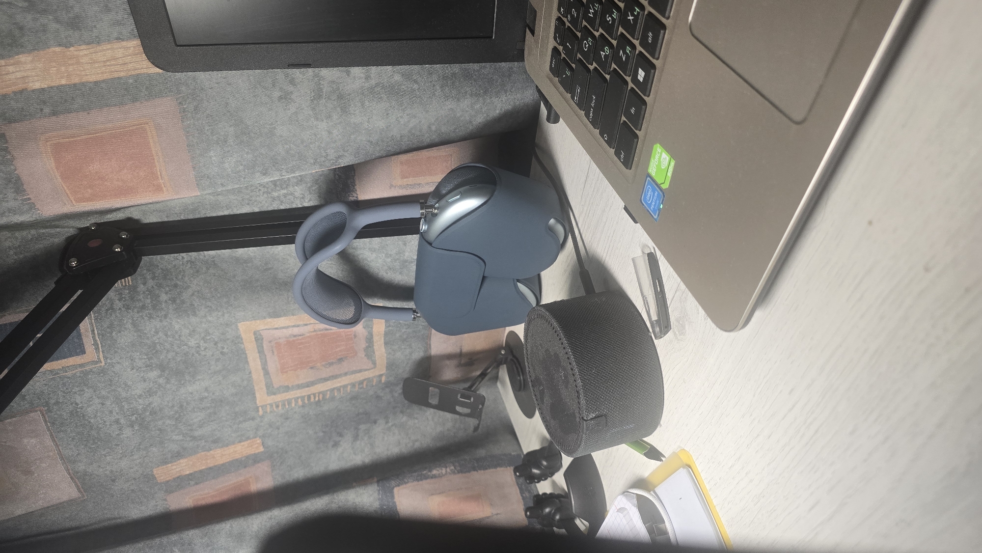 The image depicts a desk scene with various electronic devices and accessories. In the foreground, a laptop is partially visible, showing its keyboard and trackpad with stickers indicating Intel Pentium and NVIDIA GeForce graphics. To the left of the laptop, a pair of blue over-ear headphones is hanging from a black microphone stand. In front of the headphones, there is a cylindrical black speaker. Further to the left, a black phone stand is positioned behind a small black object that could be a camera lens cap or a similar accessory. A green pen rests on a yellow notepad. A small clear plastic object, possibly a USB drive or memory card case, is near the speaker. The background features a patterned curtain. There are no people visible in the image. The scene appears to be indoors, likely a home office or study. No specific landmarks or features identifiable with the city of Ukhta, Russia are present in the image.