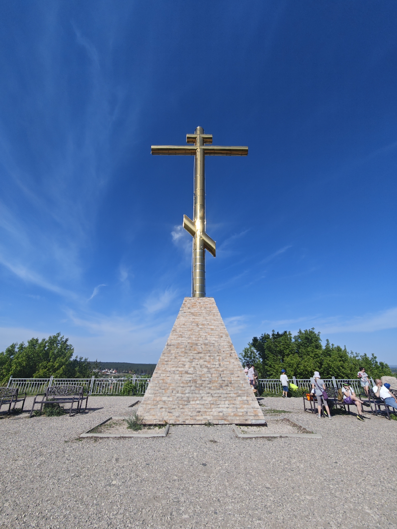 A low-angle, wide shot depicts the Memorial Sign to the Pioneers of Volzhskiy, a monument featuring a large, golden Orthodox cross erected atop a brick pyramid-shaped pedestal, set against a bright blue sky with faint, wispy clouds. The monument is situated on a paved area covered with small, light-colored stones.
To the left of the monument, several dark-colored benches face away from the camera, separated from the drop-off by a white, ornate railing. To the right of the monument, several people stand and sit on benches behind the railing.
In the background, beyond the railing, are green trees and hints of the city of Volzhskiy, Russia.