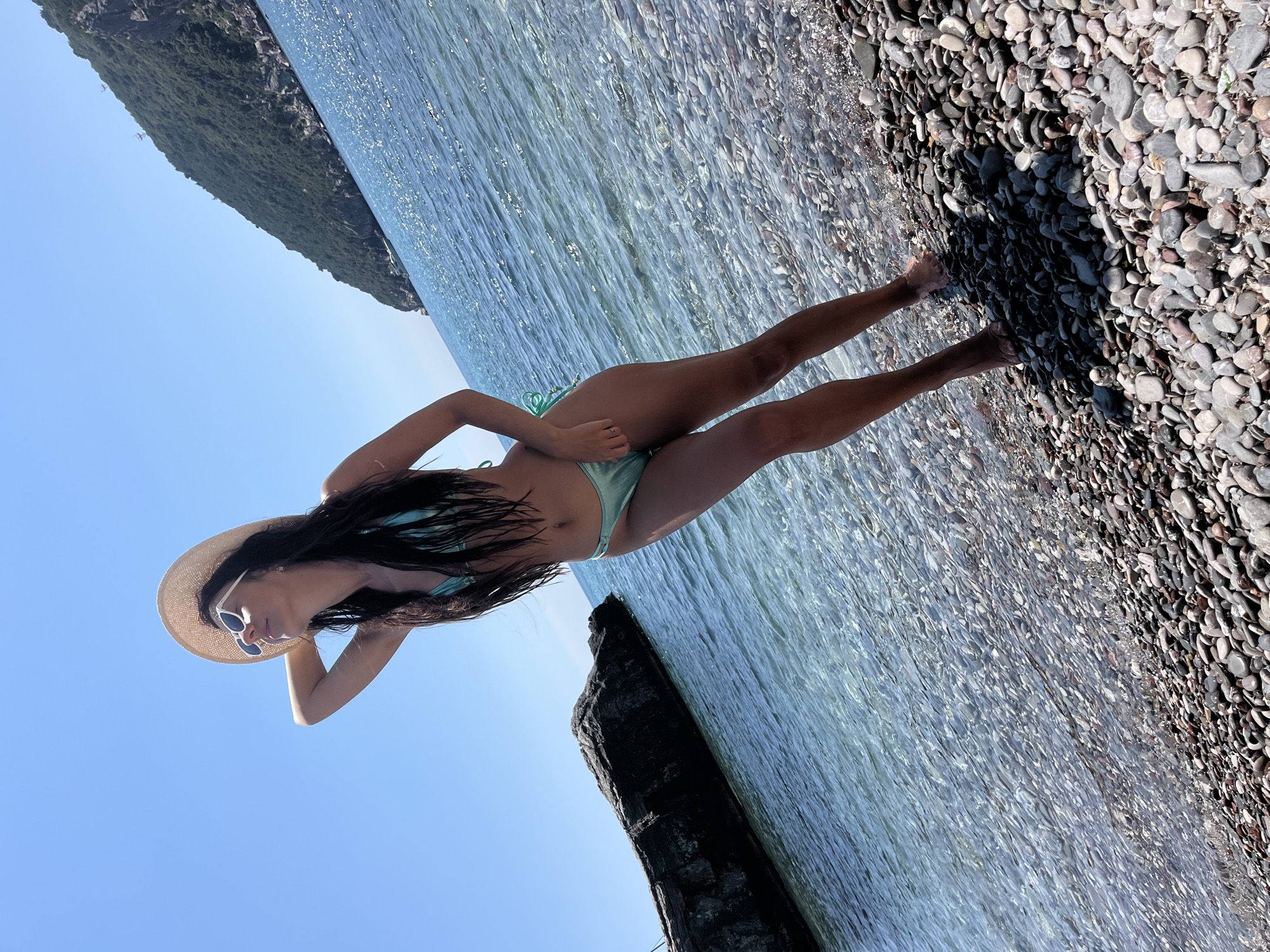 A woman in a mint green bikini and a straw hat stands on a pebble beach by the sea in Zagrađe, Montenegro. She is looking up and to the left, with her right arm raised behind her head. The water is clear and blue, with sunlight reflecting off the surface. A large dark rock is visible in the water to the left of the woman, and a tree-covered hill rises in the background. The sky is clear and blue.