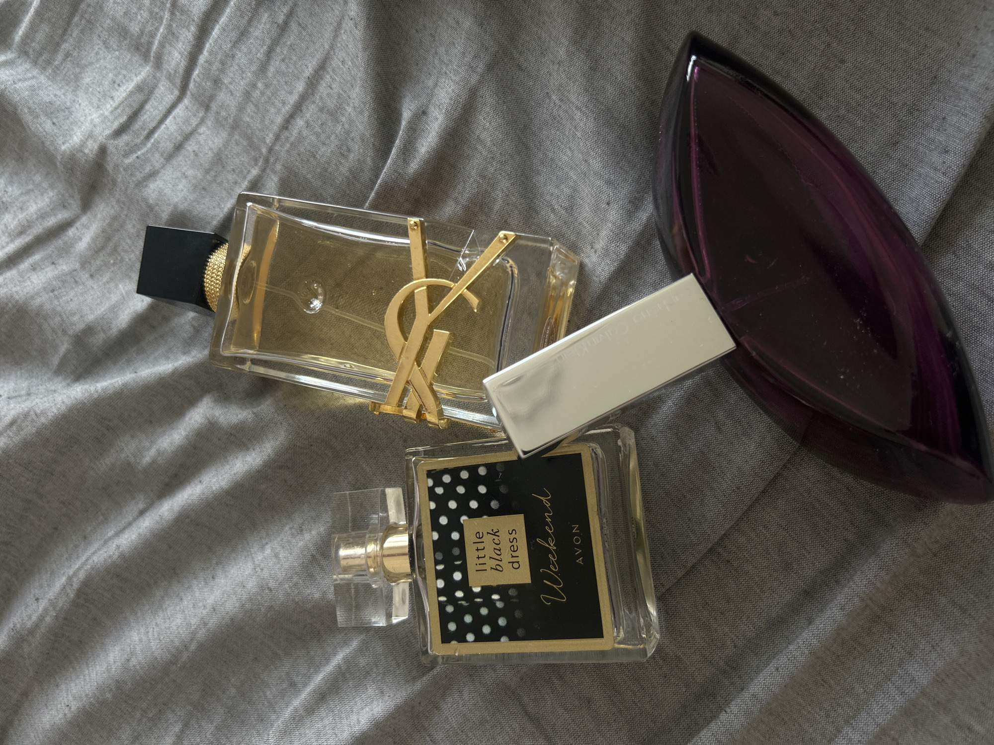 The image shows three perfume bottles on a gray, wrinkled fabric. The bottle on the upper left is a rectangular glass bottle with a gold "YSL" logo and a black cap. Next to it, slightly lower, is a rectangular glass bottle with a black label that says "little black dress Weekend AVON" in gold lettering and has white polka dots. The third bottle is a dark purple, teardrop-shaped bottle with a silver cap. The location is unknown.
