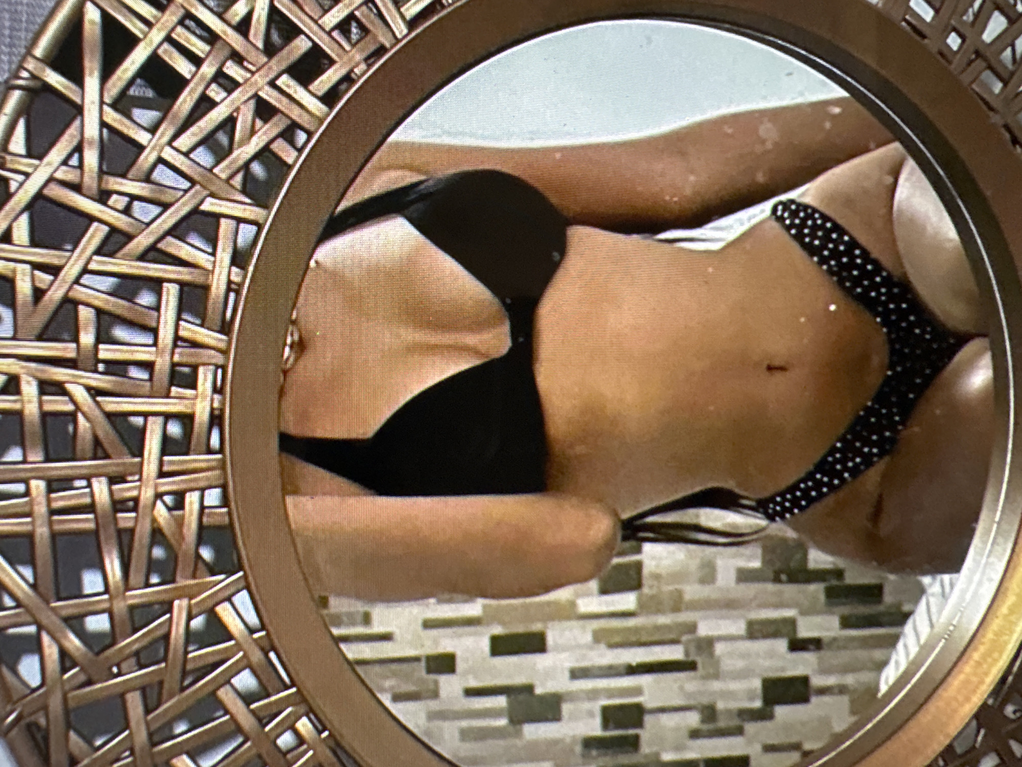 The image is a close-up photograph of a person's torso reflected in a mirror with an ornate, woven frame. The person is wearing a black bra and black bikini bottoms with white polka dots. The background reflected in the mirror appears to be a wall with a patterned tile or wallpaper and possibly a bed or furniture. The image is cropped in a way that focuses on the midsection of the person. There is no identifiable information about the location in Castelo Branco, Portugal, or any specific actions or interactions occurring. The description is limited to the visual elements present in the photograph.
