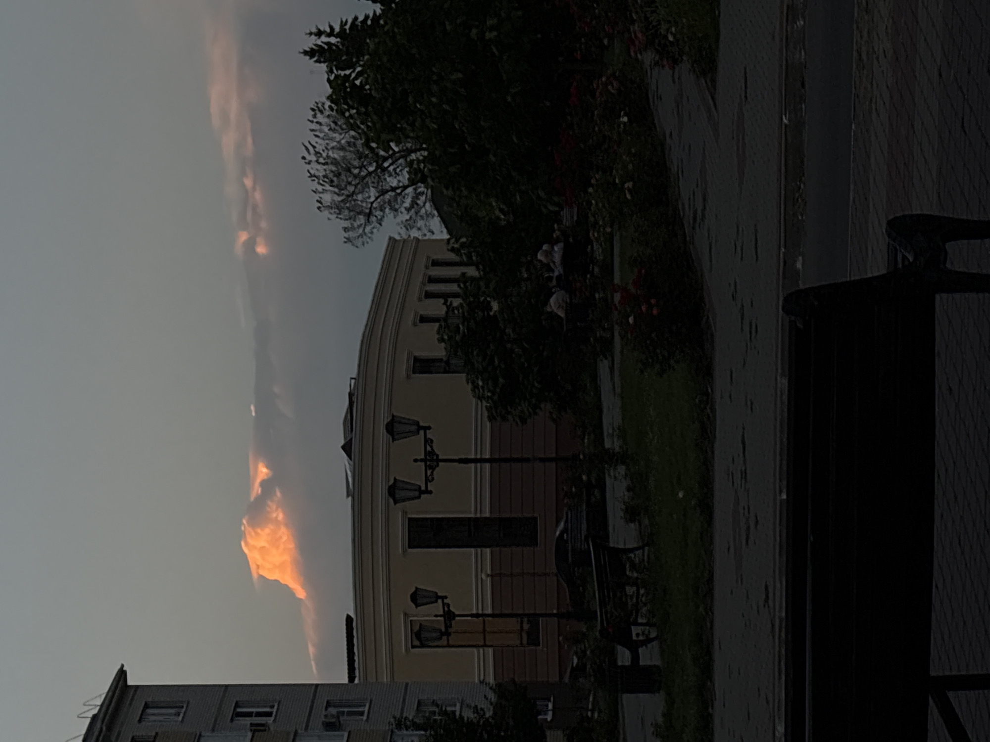 The image captures a view of a city street in Bobrov, Russia, at dusk. A tall, multi-story apartment building is visible on the left side of the frame, its windows reflecting the fading light. In the center, a light-colored, curved building with large windows and decorative trim dominates the mid-ground. A streetlamp with multiple lights stands in front of this building. To the right of the curved building, dense trees and bushes create a dark, lush backdrop. The sky above is a gradient of gray, with a single, prominent cloud illuminated by an orange glow, suggesting the setting sun. In the foreground, a dark silhouette of a park bench occupies the lower portion of the image. Scattered throughout the scene are hints of parkland, including grass, a pathway, and some flowering bushes. A group of people are seated on a bench in the middle ground, partially obscured by foliage.
