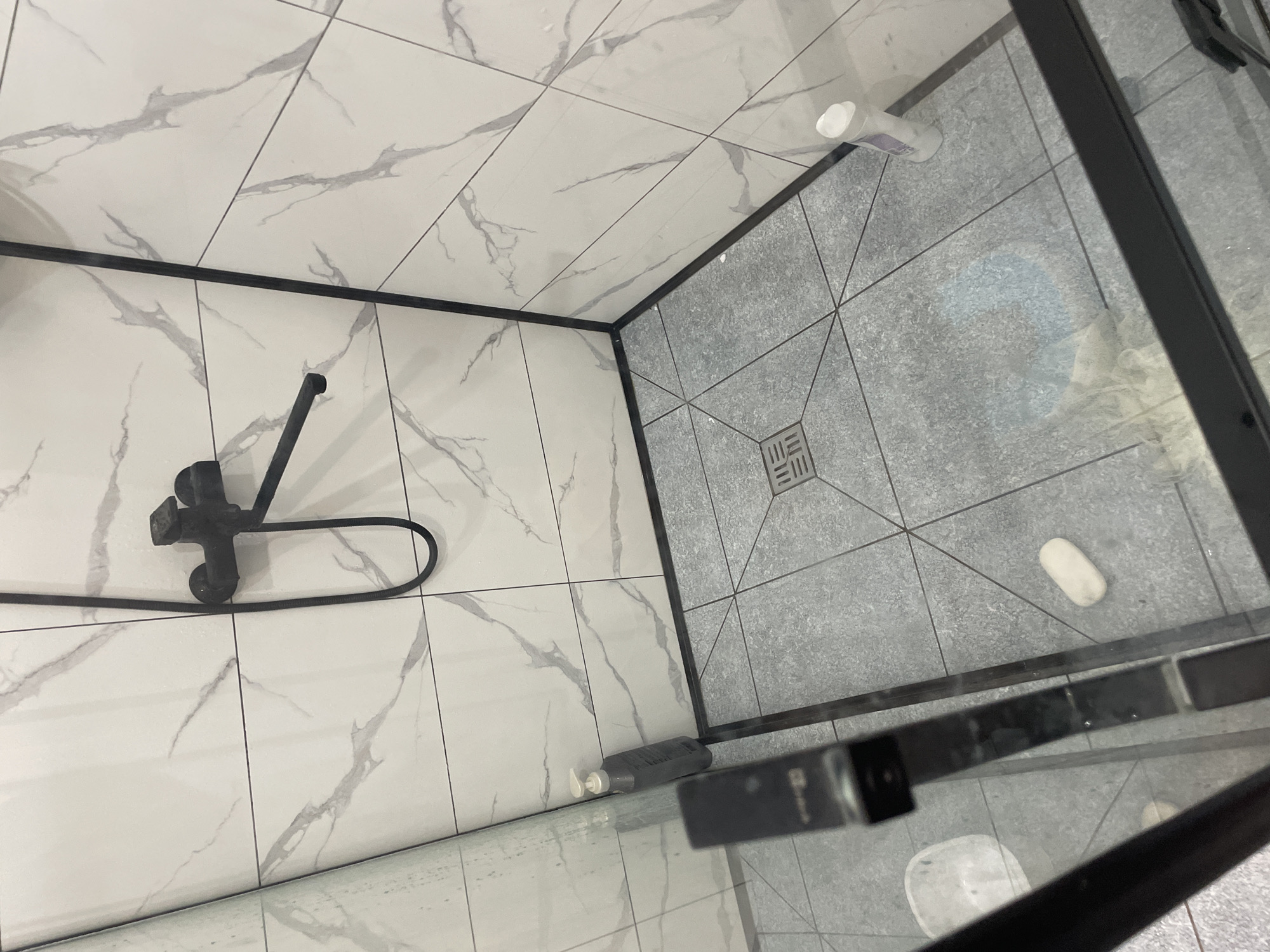 This is an indoor, eye-level, medium shot of a modern shower stall. The stall features white marble-patterned tiles on the walls and gray square tiles on the floor, with a black showerhead and faucet fixture. A dark gray soap dispenser is positioned on the left wall near the showerhead. On the floor, a white oval object, possibly a bar of soap, is visible. A white bottle, likely shampoo or conditioner, stands near the corner on the right side of the shower floor. A clear glass shower door with black framing is partially visible in the foreground, with a glimpse of a white bucket or container just outside the stall. The environment appears to be clean and recently used, with some water droplets visible on the shower walls. The location is identified as Чоң-Жар, Kyrgyzstan.
