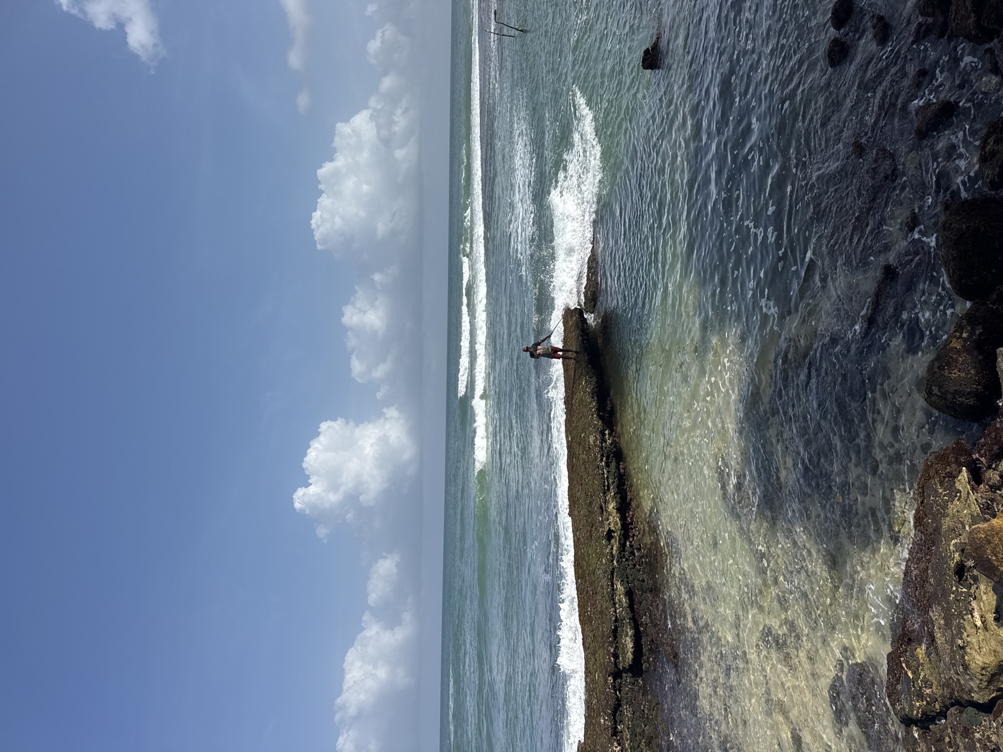 A lone fisherman stands on a rocky outcrop in the ocean off the coast of Goviyapana, Sri Lanka, casting a fishing rod into the choppy turquoise water. The fisherman, wearing red shorts and a dark shirt, is the central subject, positioned approximately in the middle of the frame. The rocky outcrop extends from the left foreground towards the center, covered in moss and seaweed, with waves crashing against its edges. Beyond the outcrop, the ocean stretches to the horizon, with distinct lines of waves. The sky above is a clear blue, punctuated by scattered white cumulus clouds. The water near the shore is clear enough to reveal the seabed and submerged rocks, while the deeper ocean appears darker. Two thin wooden poles are visible in the water to the right of the frame, likely used for fishing. The overall scene depicts a tranquil yet active fishing environment.
