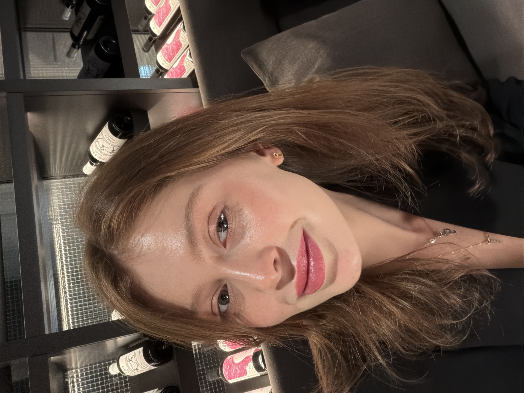 A young woman is prominently featured in a close-up, rotated portrait, appearing to be lying down while looking towards the camera. She has long, light brown hair that cascades around her and a natural, pleasant expression with a slight smile. Her skin has a dewy appearance, and she wears subtle eye makeup and pink lipstick. Visible accessories include a small, gold paperclip-shaped stud earring in her left ear and a delicate gold chain necklace with small charms, including what appears to be a stylized initial 'E' or 'L' and a tiny crystal or diamond. She is wearing a dark, possibly black or dark grey, top.
The setting appears to be indoors, likely a wine bar or restaurant, in Tbilisi, Georgia. In the background to her left, there are shelves filled with numerous wine bottles stored horizontally. The shelves are dark with a reflective, metallic grid pattern. To her right, a soft, dark grey upholstered cushion or sofa is visible, suggesting she is reclining.
Notable details on the wine bottles include labels that are partially legible. One bottle clearly shows "MARANI RKATSITELI DRY WHITE WINE" on a distinct pink label, indicating a Georgian white wine. Other bottles have labels mentioning "Château BOURGOGNE" (or BOURDEAU) with a year like "2018", and "VIN ROUGE" with years from the 1960s or 1990s, suggesting a collection of diverse wines. The lighting is ambient and somewhat dim, with direct light highlighting the woman's face.
