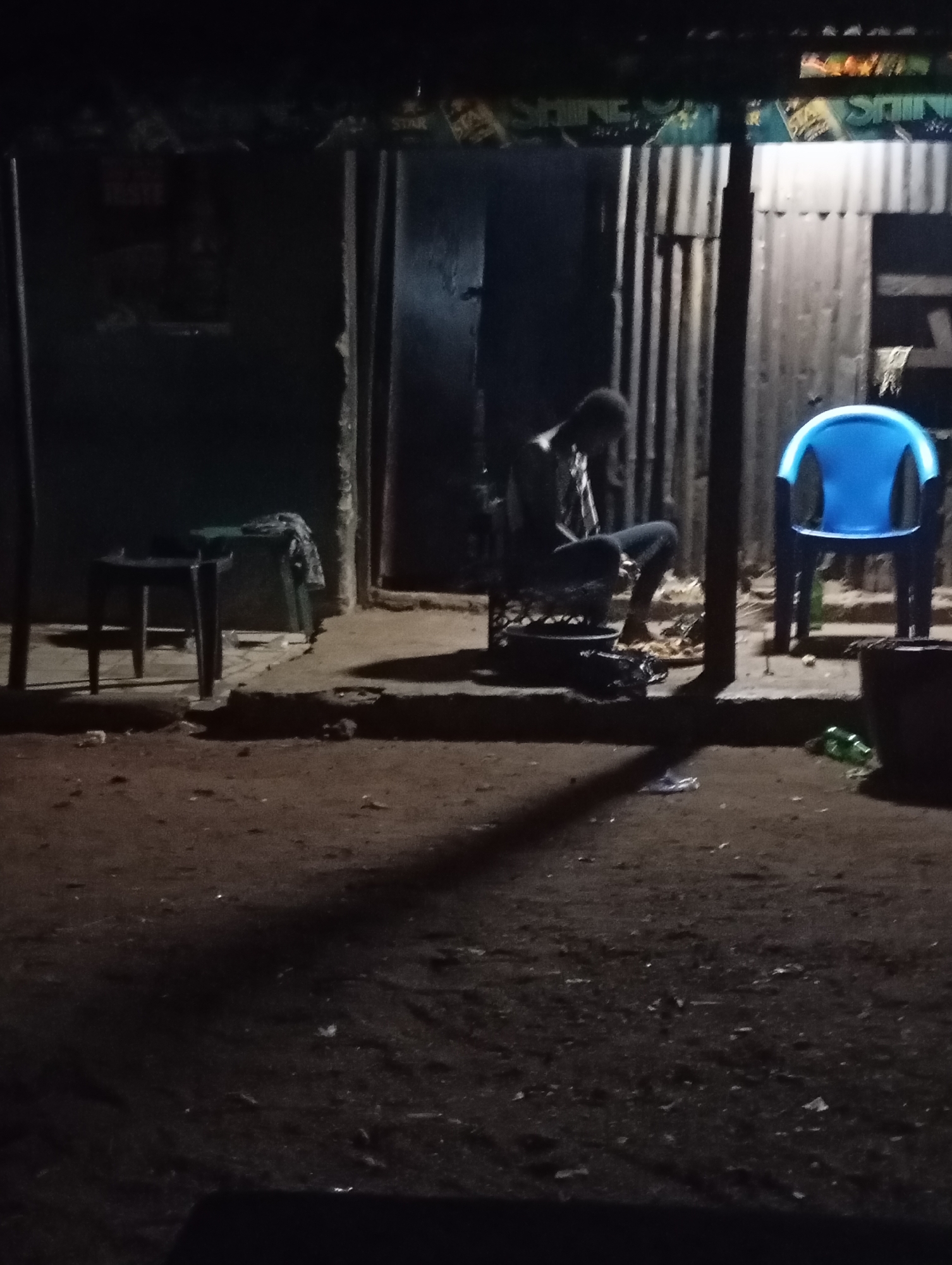 The image depicts an outdoor scene at night, likely in Shendam, Nigeria. A person is seated on a low stool or bench in front of a corrugated metal structure, appearing to be engaged in an activity on the ground before them. To their right, a bright blue plastic chair stands empty. The ground is unpaved and appears to be dirt or gravel, with scattered debris. The background shows a dimly lit structure with corrugated metal siding and a partially visible sign that reads "SHINE". The overall impression is one of a humble, possibly open-air establishment, illuminated by ambient light and possibly some artificial lighting that is not directly visible.