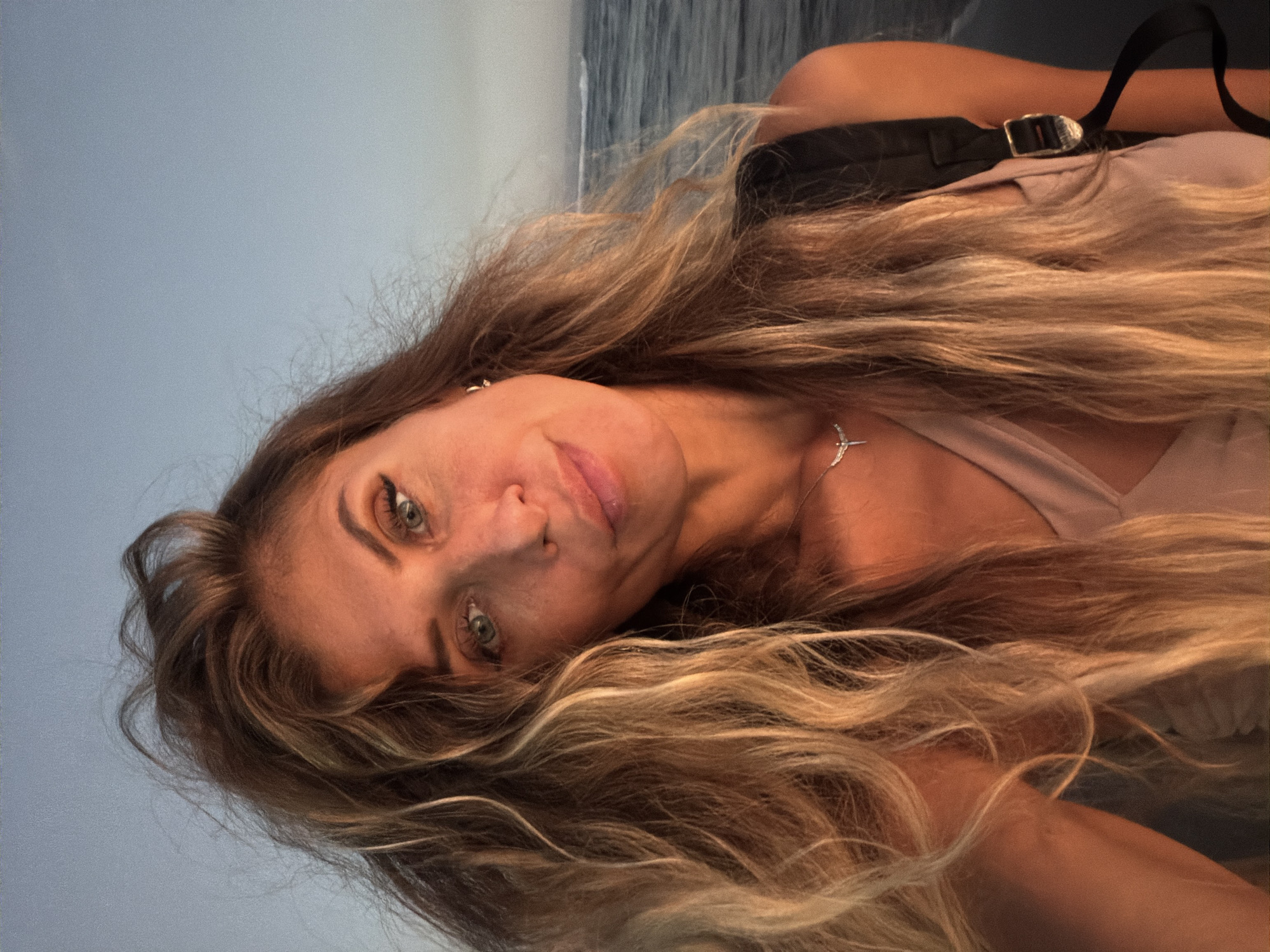 A selfie is taken by a woman with long, wavy blonde hair and blue eyes. She is wearing a light-colored top and a silver necklace. She has light makeup on, including eyeliner and mascara. The woman is looking towards the camera with a slight smile. Behind her, there is the ocean with some waves and a clear sky. The image appears to have been taken at sunset, with warm lighting casting a glow on the woman's face and hair. The location is the coast of North Kuta, Indonesia.