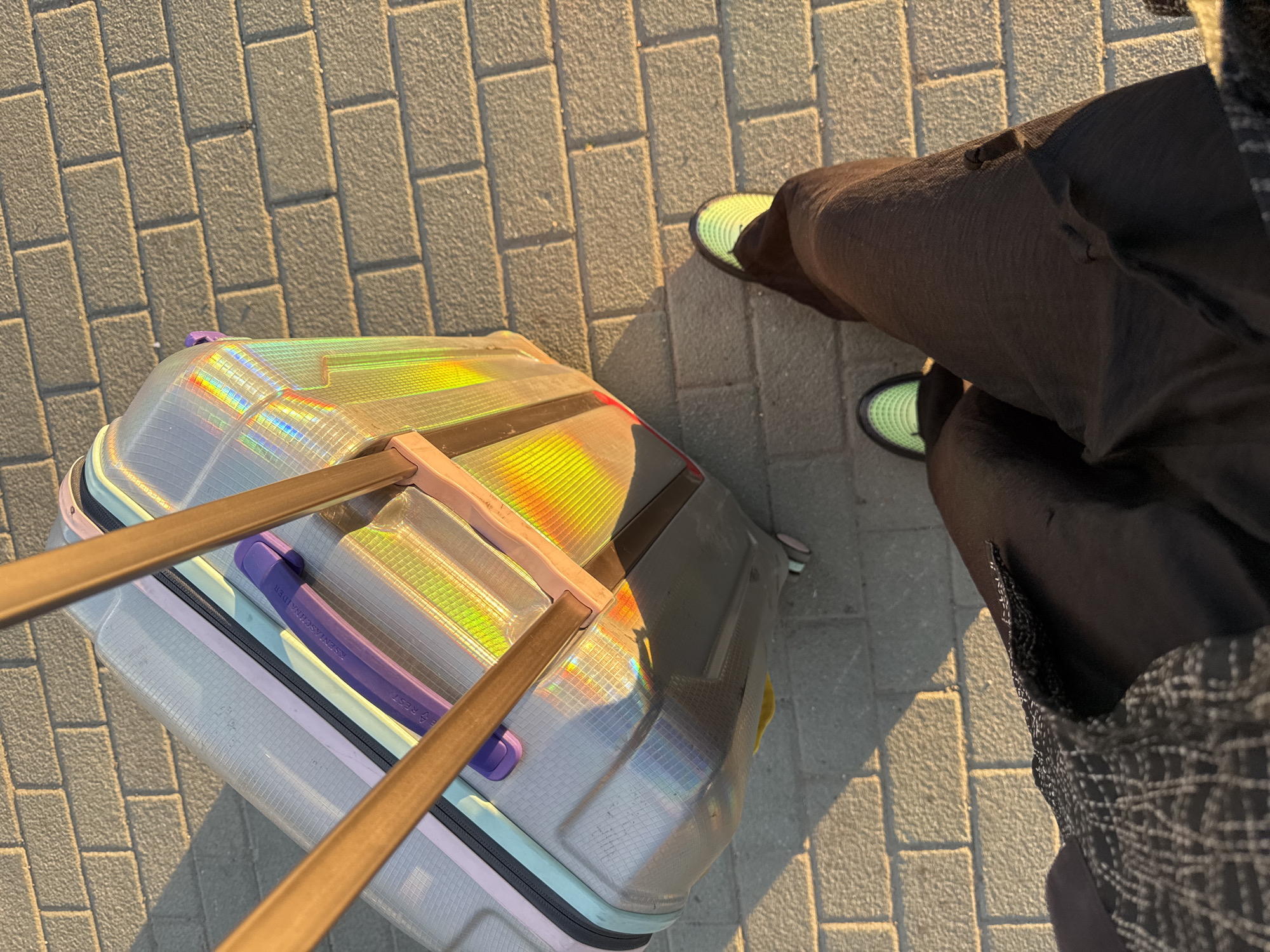 A person stands on a paved surface, next to a suitcase. The pavement is constructed of light gray rectangular bricks, laid in a running bond pattern. The suitcase is positioned on the left side of the frame, displaying a holographic sheen on its surface that reflects rainbow colors. The suitcase features a retractable handle and bands of different colors. The person standing beside the suitcase is wearing dark-colored trousers and light green sneakers. The overall perspective is from above, looking down. There is no information within this image to determine the location in the city of Moscow, Russia.