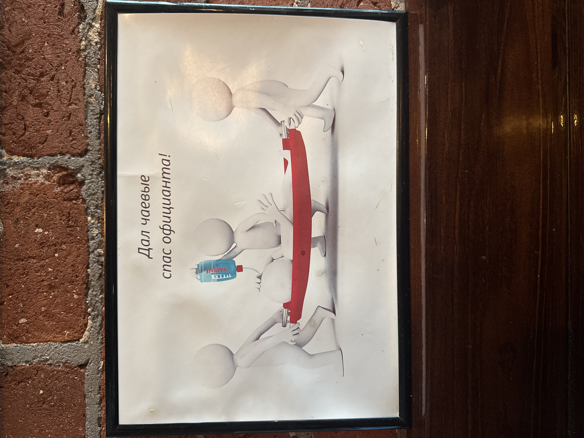 The image shows a framed sign hanging on a brick wall next to a wooden door. The sign depicts a cartoonish scene of three white stick figures working together to rescue a fourth stick figure, who is holding a blue bottle labeled "ЧАЕВЫЕ" (Chayevye), which translates to "Tips". The rescuing figures are using a red bar as a lever. The text on the sign reads "Дал чаевые - спас официанта!" (Dal chayevye - spas ofitsiant!), meaning "Give a tip - save the waiter!". This suggests the image is located in a restaurant or cafe in Moscow, Russia. The sign is a humorous appeal to customers to leave tips for the wait staff.