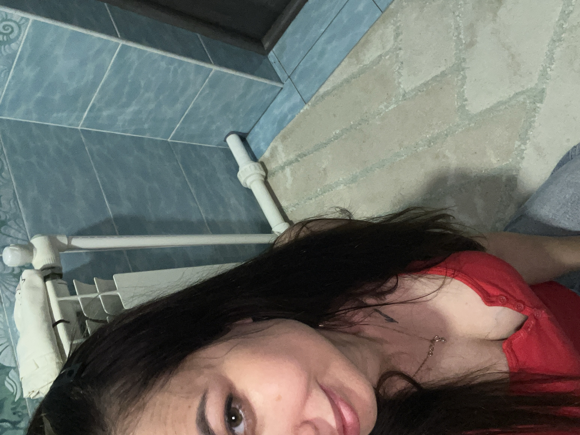 A woman with long, dark hair is pictured from a high angle in what appears to be a bathroom. She is wearing a red top and a necklace. The background consists of blue tiled walls, a white radiator, a white pipe, and a patterned floor mat. The lighting is dim.