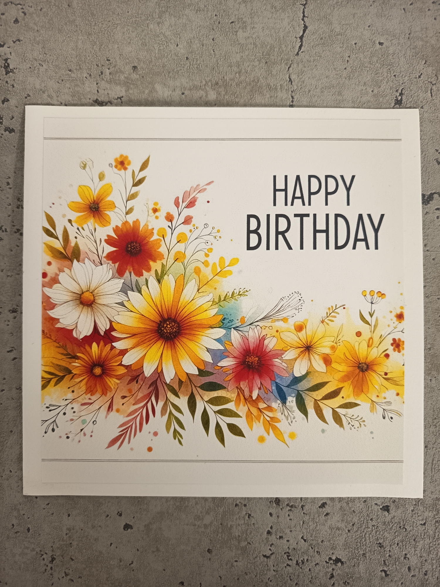 The image shows a birthday card with a floral design. The card has a white background with a border line at the top and bottom. The left and lower portion of the card features an arrangement of watercolor-style flowers in shades of yellow, orange, red, and white with green leaves and delicate accents. The words "HAPPY BIRTHDAY" are printed in large, bold, black capital letters in the upper right corner of the card. The card is placed on a gray, textured surface. The location of Wangen bei Olten, Switzerland is not determinable from the content of the picture.