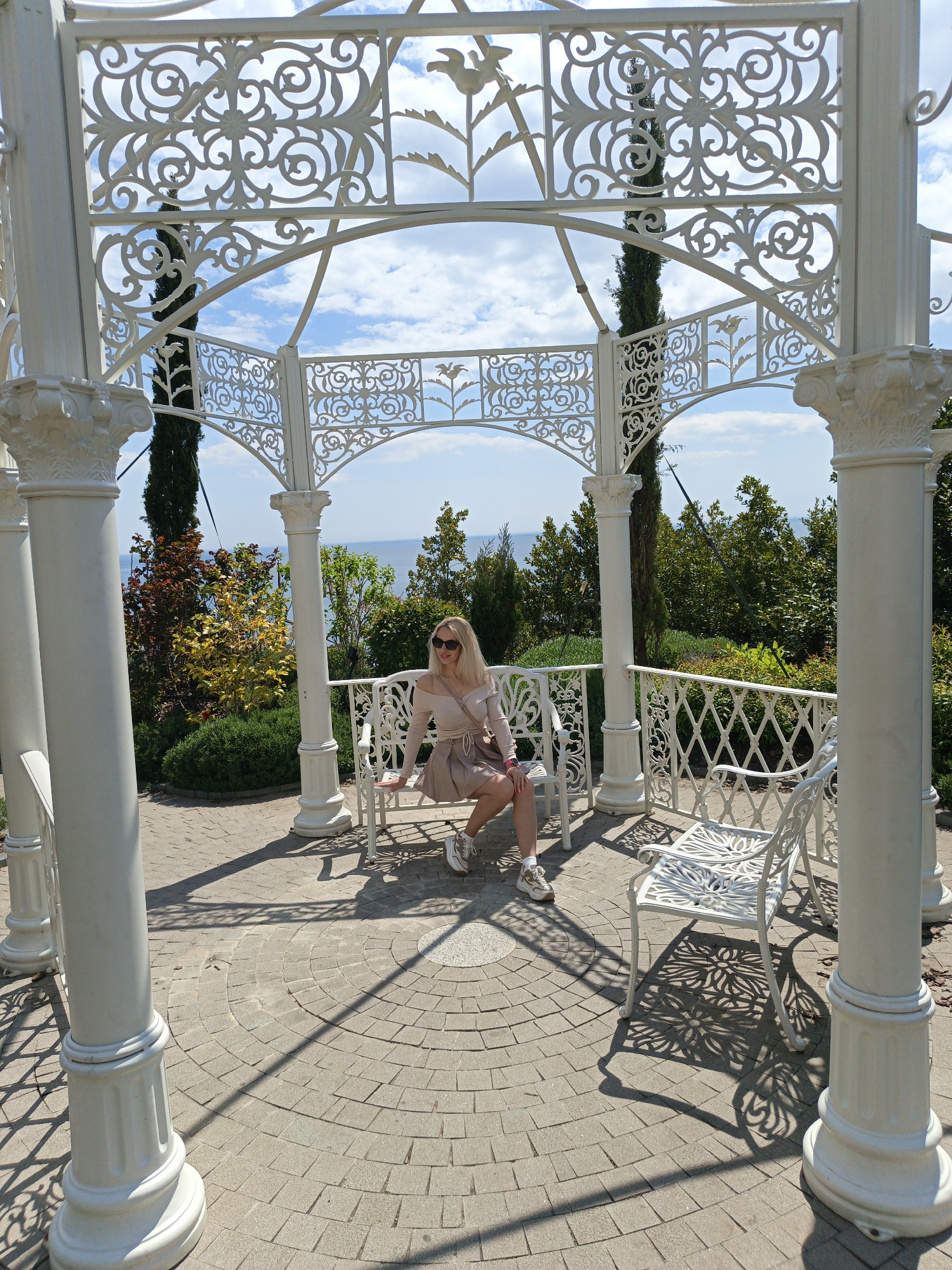 A full, eye-level shot depicts a woman seated on a white metal bench within an ornate white gazebo structure, situated in Unknown, Unknown. The gazebo features decorative white metal lattice designs and white columns.
The woman is wearing sunglasses, a light pink top, and a matching skirt. She has a purse slung over her shoulder and wears sneakers. She is seated on a white metal bench, facing the camera. Another identical, empty bench is to her right.
The gazebo is surrounded by lush greenery, including trees and bushes. In the distance, a sliver of blue ocean is visible. The sky is partly cloudy, with patches of blue peeking through. The gazebo floor is made of patterned brick, casting shadows from the structure. The scene is well-lit, indicating a sunny day.