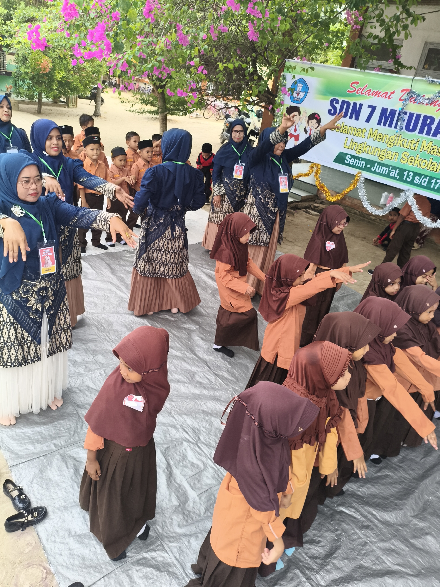 In the city of Baratpayaitek, Indonesia, a group of young girls in hijabs and school uniforms participate in an outdoor event. The scene takes place on a sandy surface covered with a grey tarp. In the background, a banner reads "Selamat Tasyah" and "SDN 7 MEURASA," suggesting a school event at SDN 7 Meurasa. The girls, dressed in brown and orange attire, are positioned in rows, some with their hands extended forward. Two women in navy blue attire, possibly teachers, are also present, one interacting with the girls. Pink bougainvillea flowers are visible on trees overhead, adding a touch of color to the environment. Shoes, including a pair of black Mary Jane shoes, are visible on the tarp.