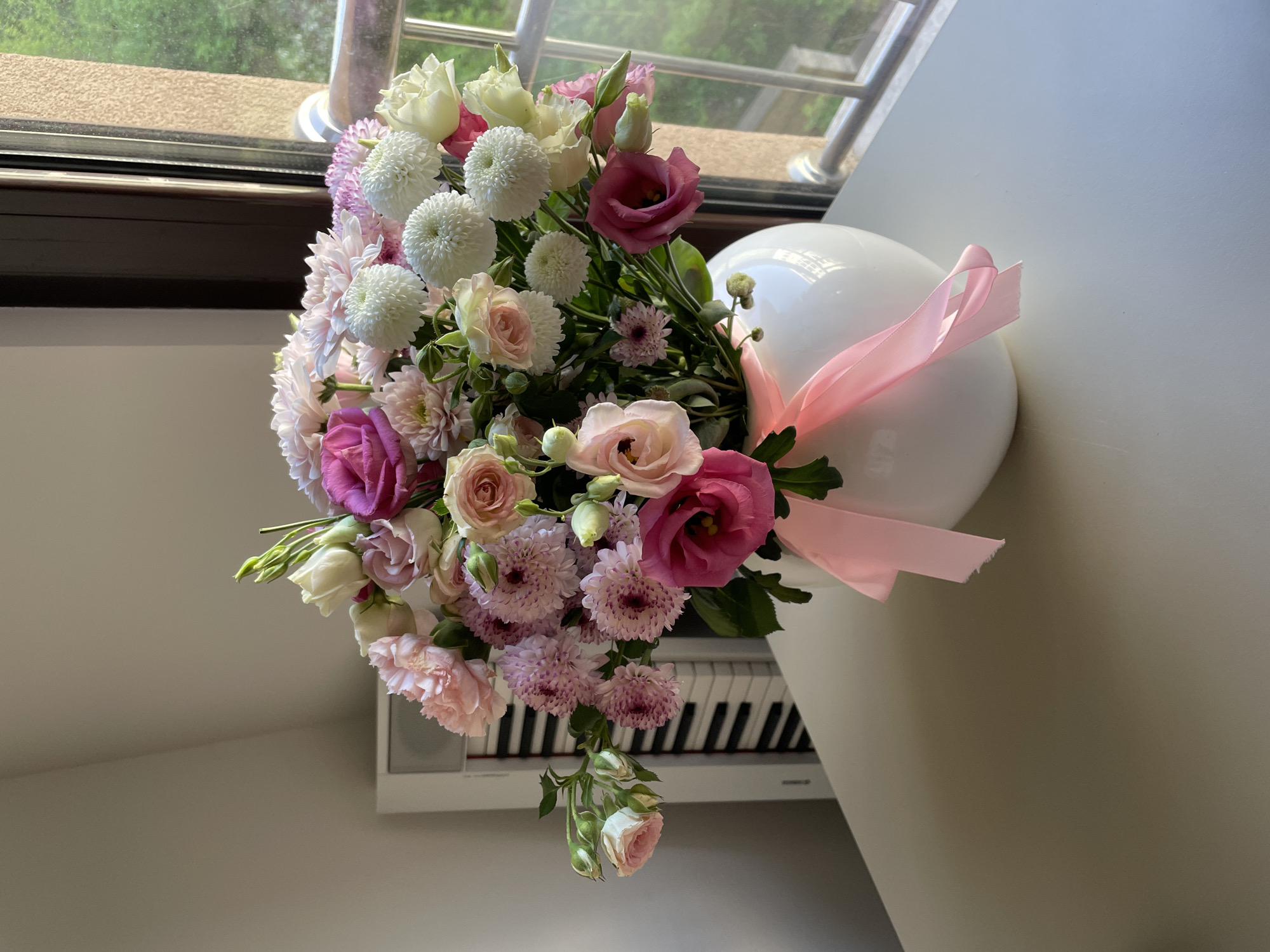 A high-angle shot shows a bouquet of flowers in shades of pink, white, and purple, positioned on a white hemispherical container with a pink ribbon attached. The bouquet sits next to a white keyboard. The background includes a window with a view of trees and foliage, and the scene is well-lit. The location is potentially in a residential setting, possibly in a room in Boreti, Montenegro.