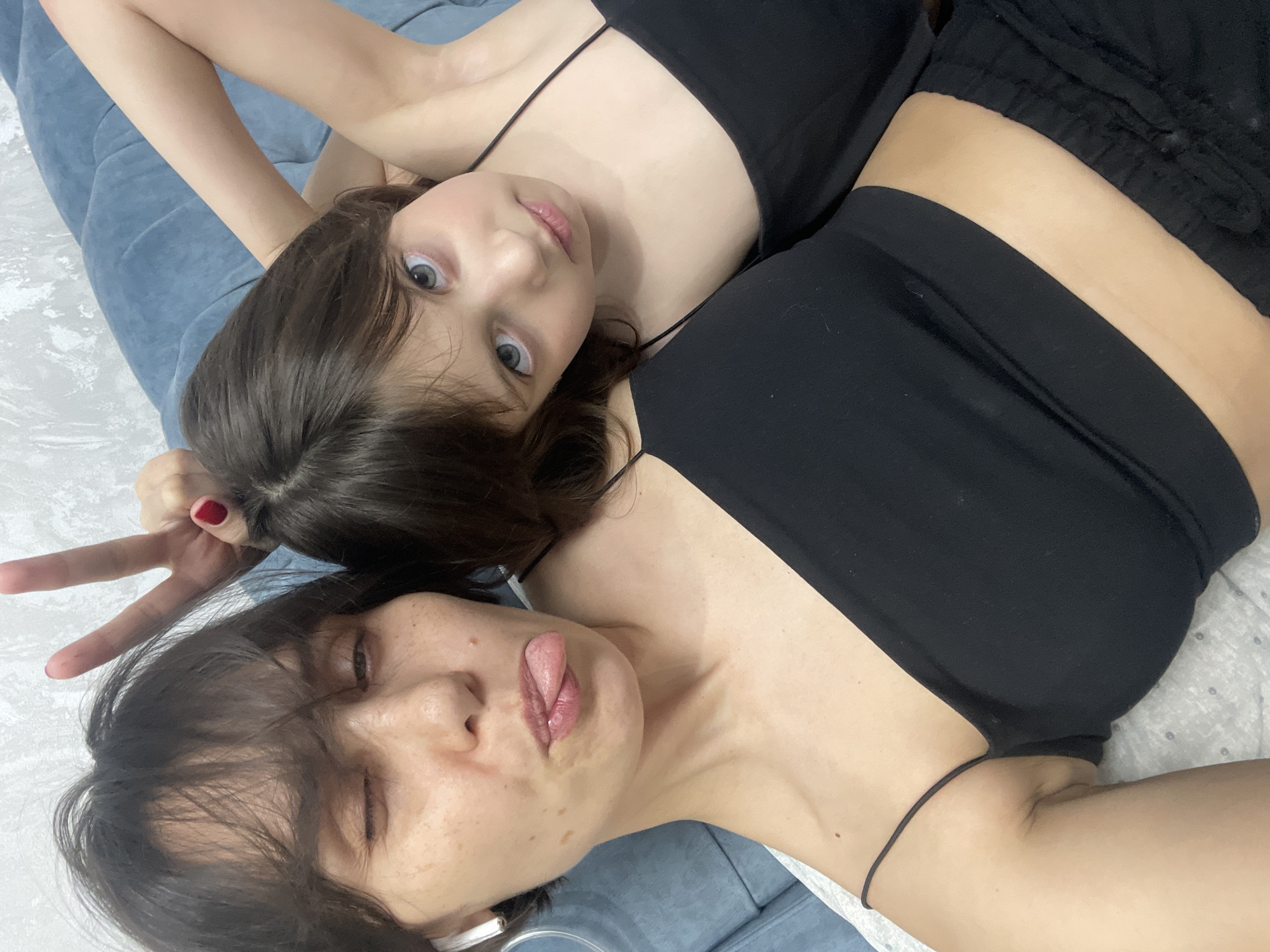 Two women are lying down. One woman is facing the camera with wide eyes and making a peace sign with her fingers. The other woman is sticking out her tongue and winking. Both are wearing black tops. The women are lying on a blue and white patterned surface.
The image doesn't have enough information to determine the location in the city of Izhevsk, Russia.