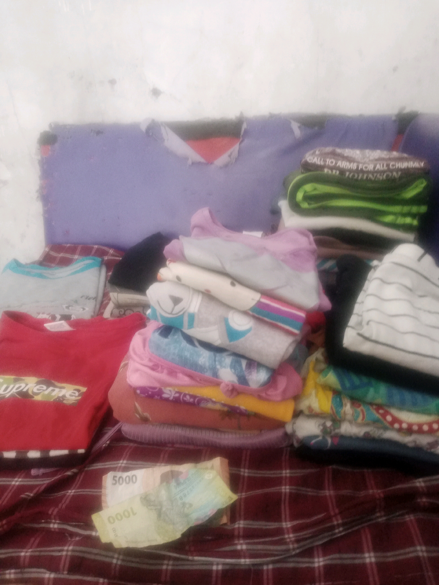 The image depicts a pile of folded clothes in a room with a light-colored wall. The clothes include a red shirt with the word "Supreme" on it, a light blue shirt, a green shirt with text, and several other multi-colored garments. Two pieces of currency, one with the denomination "5000" and another with "1000", are placed on top of a checkered fabric. The scene appears to be indoors, in Pandeyan, Indonesia, though no specific landmarks are visible. The lighting is dim, suggesting it might be a domestic setting. There are no people visible in the image.