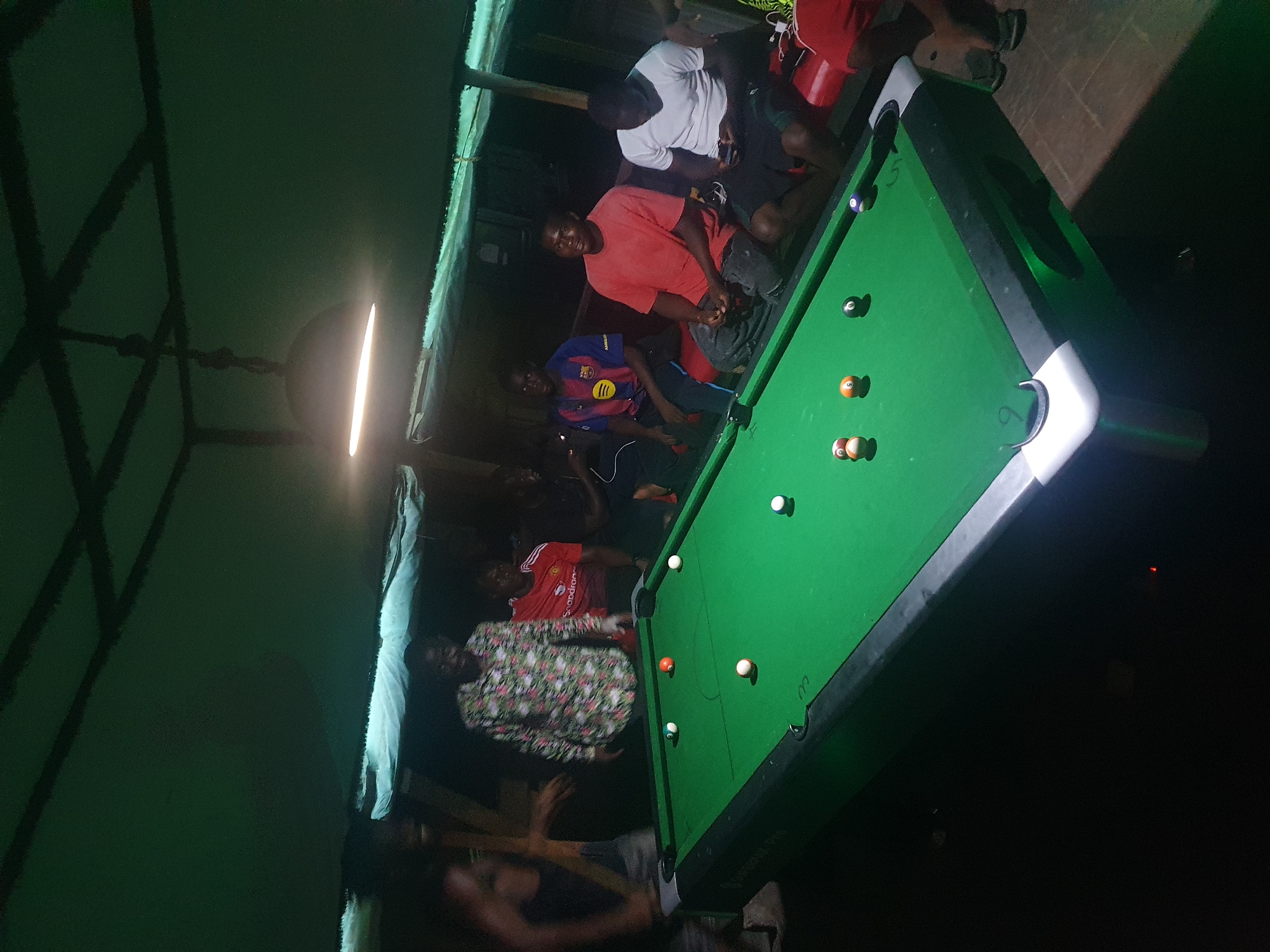 A dimly lit recreational space shows a pool table surrounded by people in what appears to be Lokoja, Nigeria. The pool table is green, with several pool balls positioned on it. The room is illuminated by a single overhead light. Several men are gathered around the pool table, some seated and some standing. A man wearing a white shirt, a man wearing a red shirt, and a man wearing a Barcelona jersey are visible near the pool table. Another man is seen wearing a floral-patterned shirt. The background includes structural elements indicating the location may be an open-air or semi-enclosed space. The overall atmosphere suggests a casual gathering.