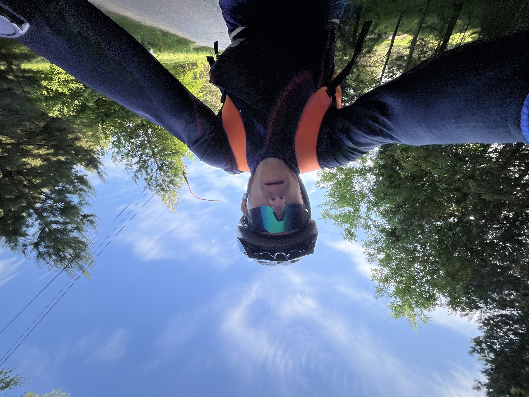 A selfie photograph looking upward shows a person wearing a black cycling helmet and reflective sunglasses. The person is wearing a black long-sleeved jacket with orange detailing and a backpack. The background features a blue sky with streaky clouds and trees with green foliage on either side. A section of pavement is visible at the top of the frame. There are electrical wires present in the upper left quadrant of the photograph. The location is likely a bike path in an area with trees, however, it is not possible to confirm the specific location in the city of Chaikovsky, Russia, without additional context.