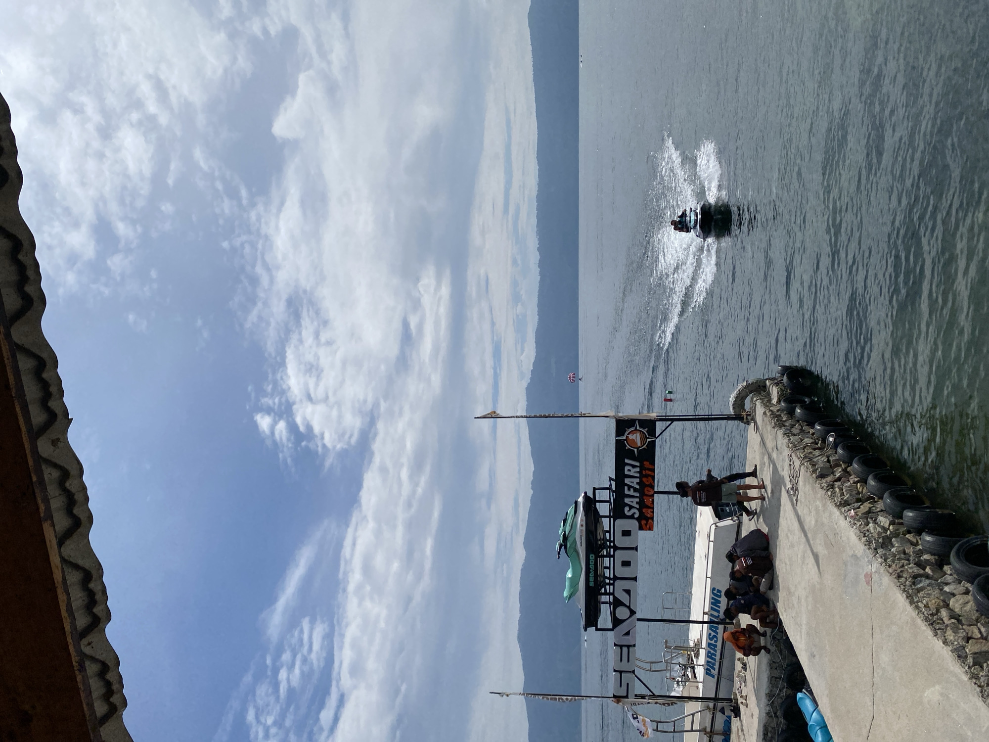 A jet ski is being ridden on a body of water near a pier in Tuk Tuk, Indonesia. The pier is made of concrete and lined with tires. A boat is docked at the pier, with a jet ski on a display stand above it. Several people are sitting on the pier, and one person is standing. A sign above the boat reads "SEA-DOO SAFARI SAMOSIR". The sky is cloudy, and mountains are visible in the background.