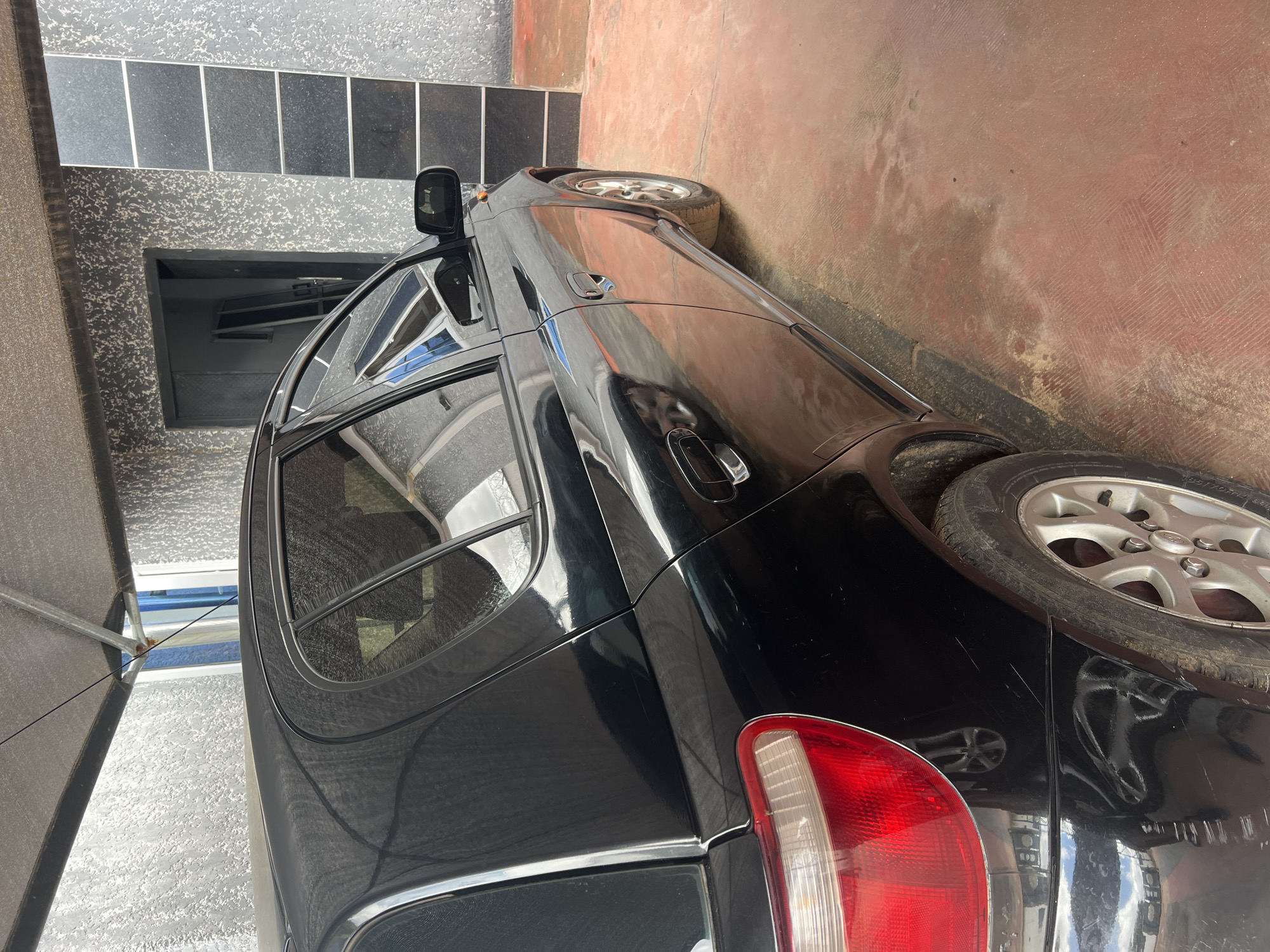 The image shows a black car parked under a grey canopy. The car is the main subject, with its side facing the viewer. The car appears to be a hatchback with tinted windows and a rear spoiler. The left rear tire is visible, and it is a silver alloy wheel. The car is parked on a reddish-brown, tiled surface. To the left of the car, a textured grey wall with dark rectangular panels is visible. The lighting suggests it is daytime, possibly outdoors, as indicated by the shadows and the natural light illuminating the scene. The overall impression is that of a car parked in a residential or commercial area. There is no discernible text in the image.