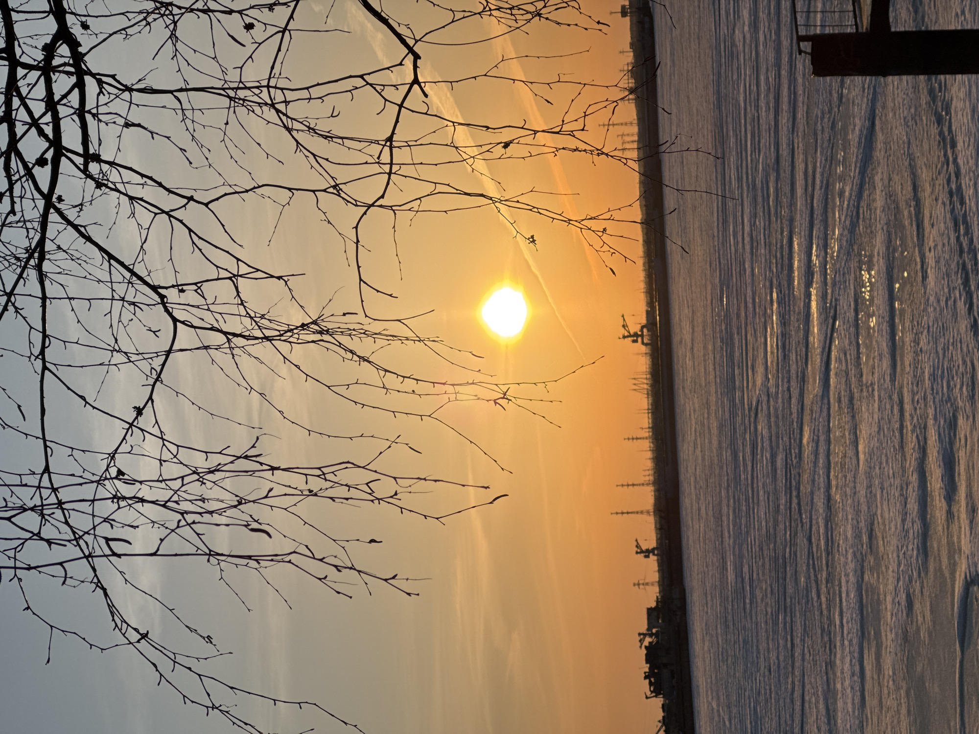 The image shows a sunset over the Kama River in Chaikovsky, Russia. Bare tree branches are silhouetted in the foreground. The sun is a bright orange orb in the center of the sky. The river is partially frozen, with a visible ice sheet. Power lines and industrial structures are faintly visible in the distance along the riverbank. A metal structure is visible in the top right.