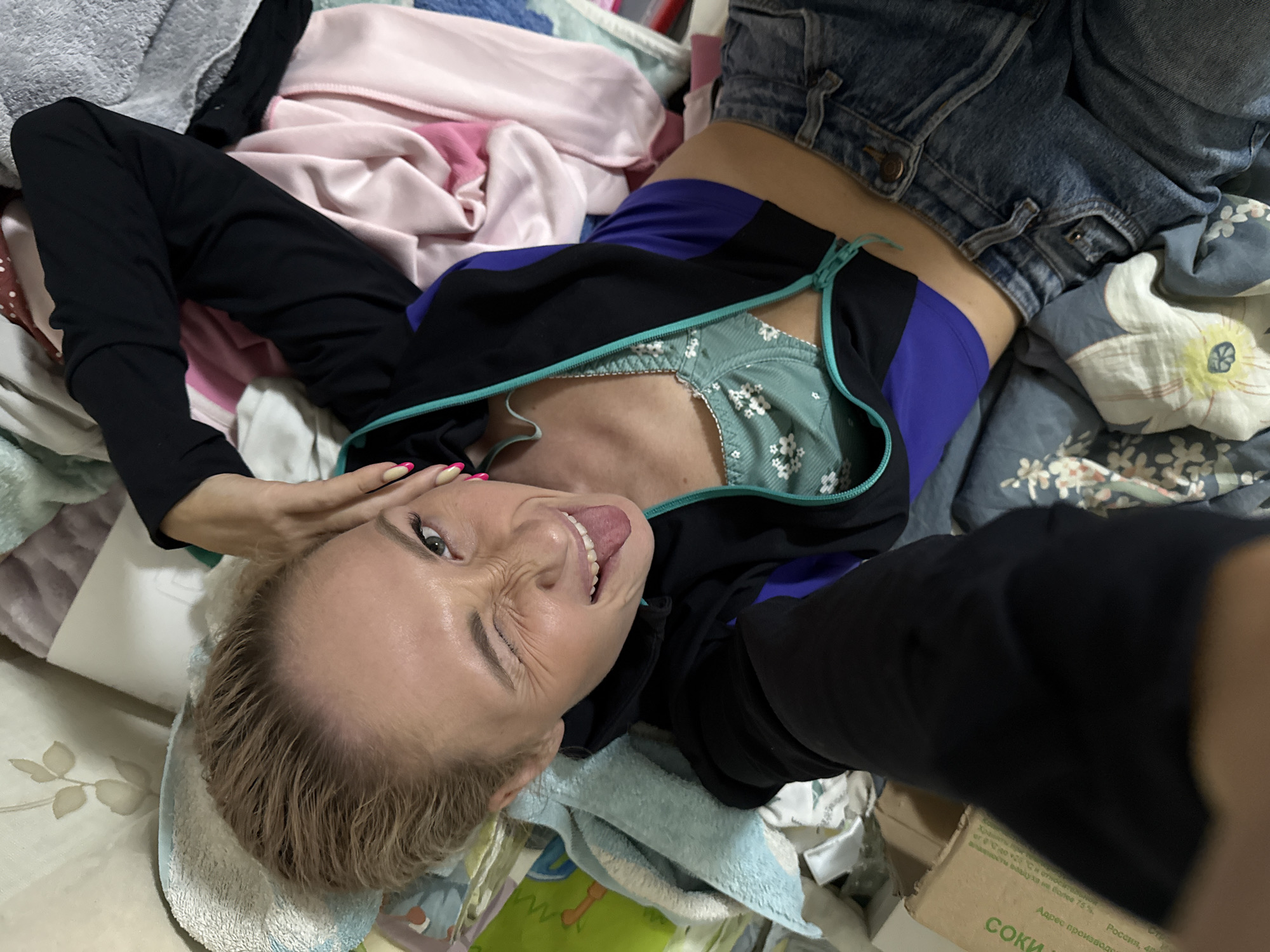 A woman is lying on a bed, taking a selfie. She is wearing denim shorts, a black and purple jacket, and a green bra with a floral pattern. She is sticking her tongue out and winking at the camera. The bed is covered in clothes and other items. A box with "Соки" printed on it is also visible in the lower right corner of the image. The box also contains Russian text and mentions "Россия, 48," which could be a reference to Chaikovsky, Russia.