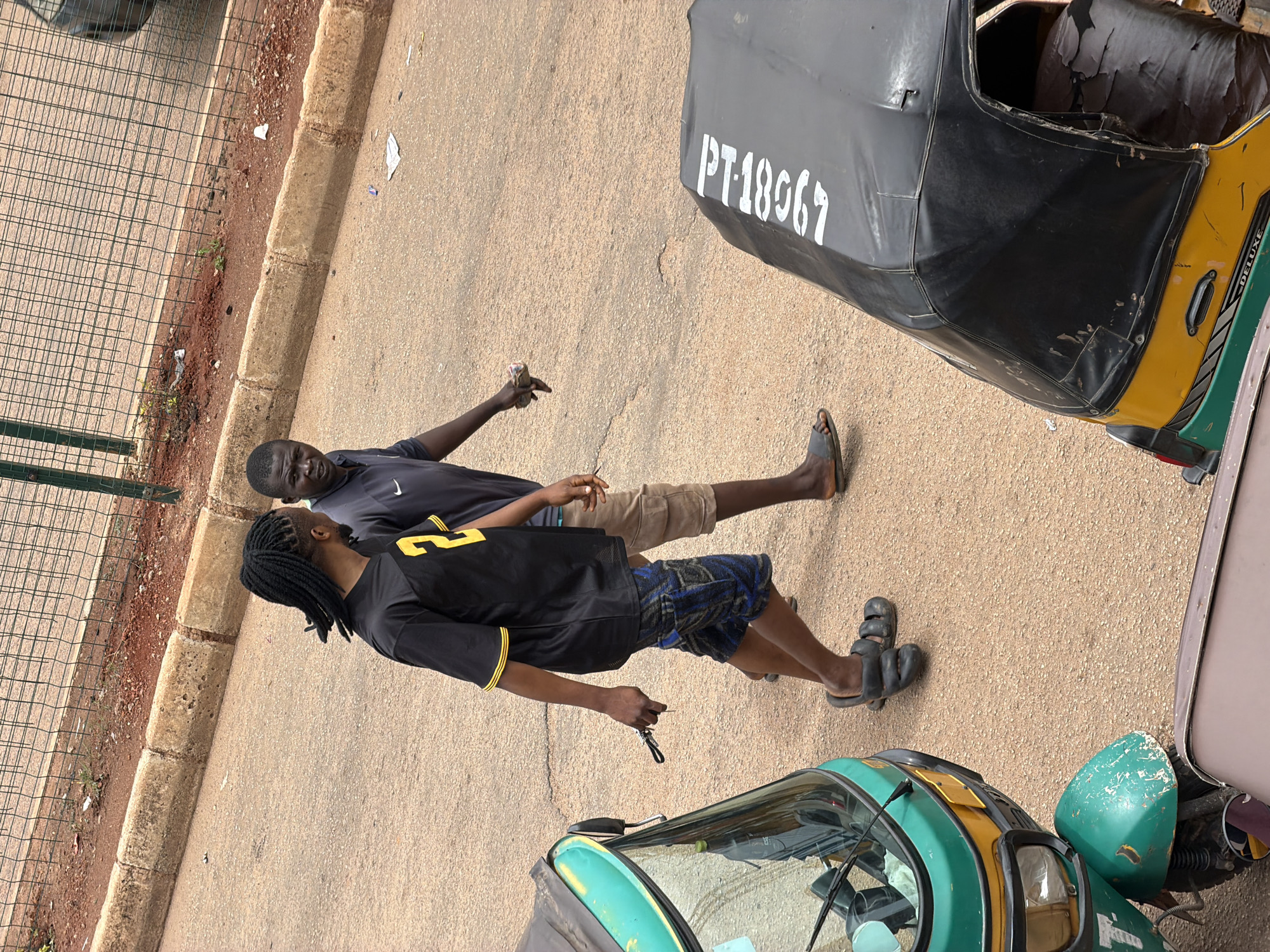 Two men are walking on a paved road in Bukuru, Nigeria. The man on the left, with braided hair, is wearing a black t-shirt with yellow trim and blue patterned shorts. He is holding a set of keys in his right hand. The man on the right is wearing a dark blue t-shirt and light brown cargo shorts. He is looking towards the camera. In the foreground, a green and yellow auto-rickshaw is parked on the left, and a black and yellow auto-rickshaw with the registration "PT-18G67" is parked on the right. The background shows a metal fence and a dirt embankment.