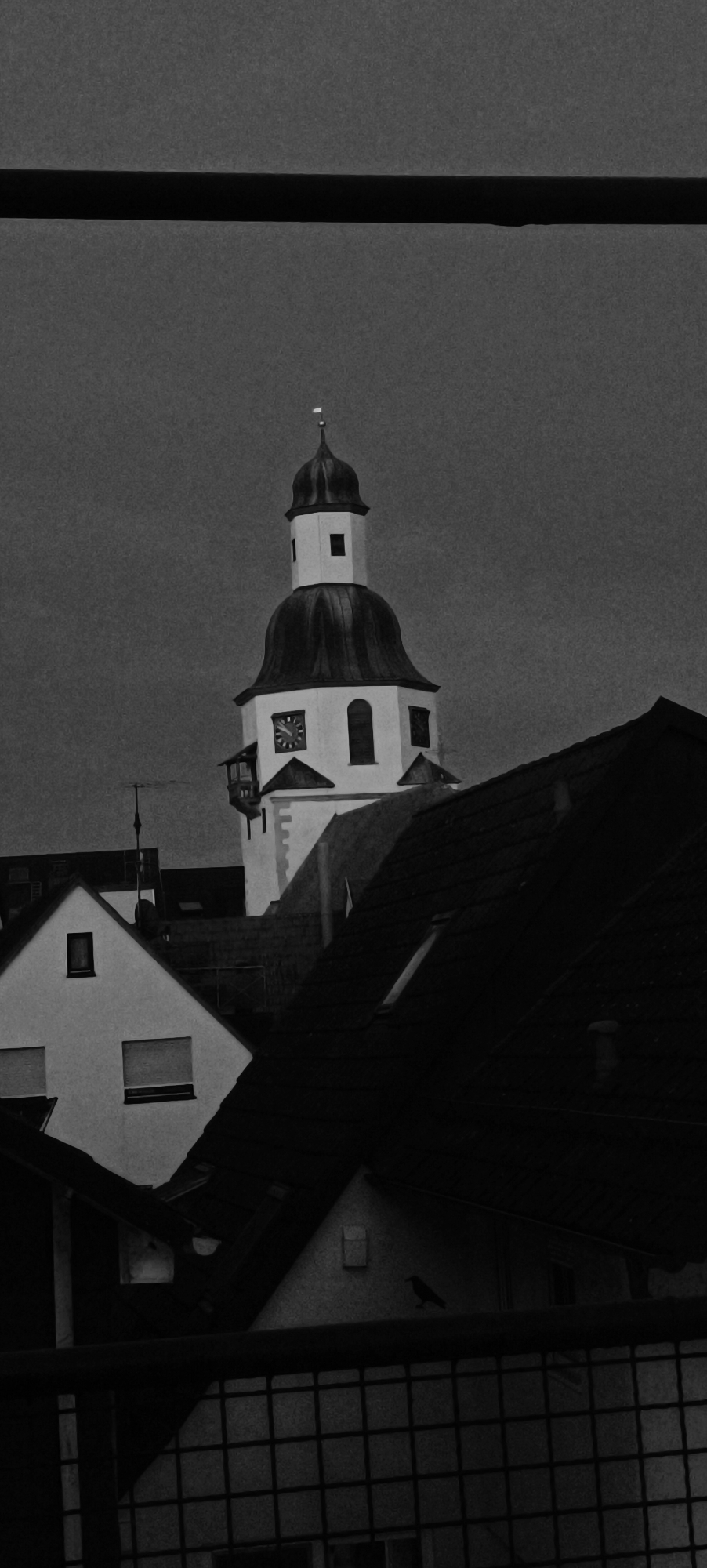 The image is a black and white photograph depicting a view of rooftops in Winnenden, Germany, with a prominent church tower in the background. The sky is overcast and dark. In the foreground, a black horizontal bar runs across the top of the frame, obscuring the top of the scene. Below this bar, rooftops of various houses are visible, some with dark tiled roofs and others with lighter-colored walls. A small black bird is perched on the angled roof of a house on the right side of the frame.
The focal point of the image is a white church tower with a dark, domed roof topped by a small spire and a white flag. The tower features a clock face, arched windows, and a small balcony-like structure. The clock face on the tower shows the time as approximately 2:35. The tower is situated behind the foreground rooftops.
The overall impression is one of a somewhat somber, urban scene, possibly taken during dusk or on a cloudy day. The photograph appears to have a grainy texture.